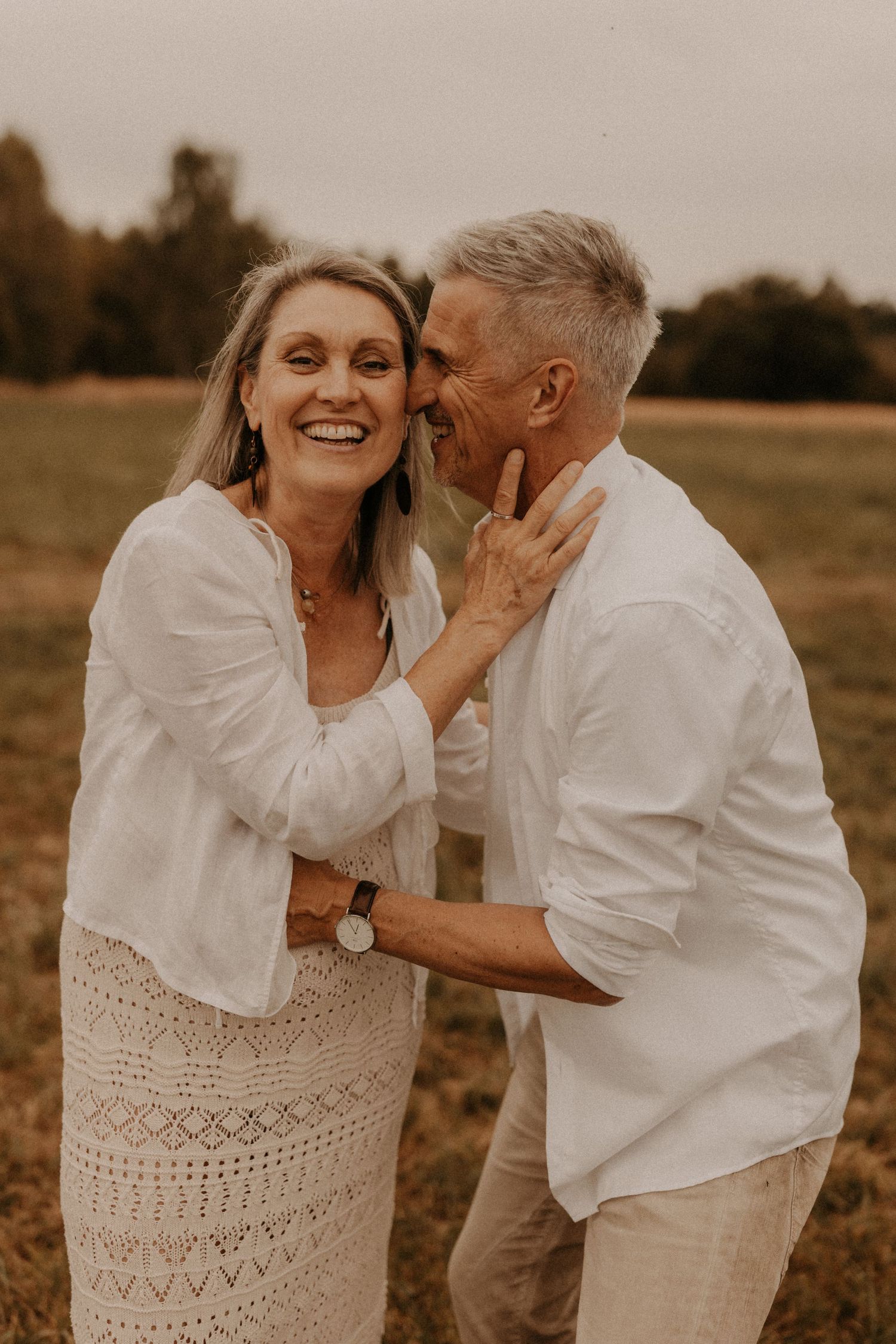 A joyful mature couple in white outfits share a tender moment laughing together in an open field.