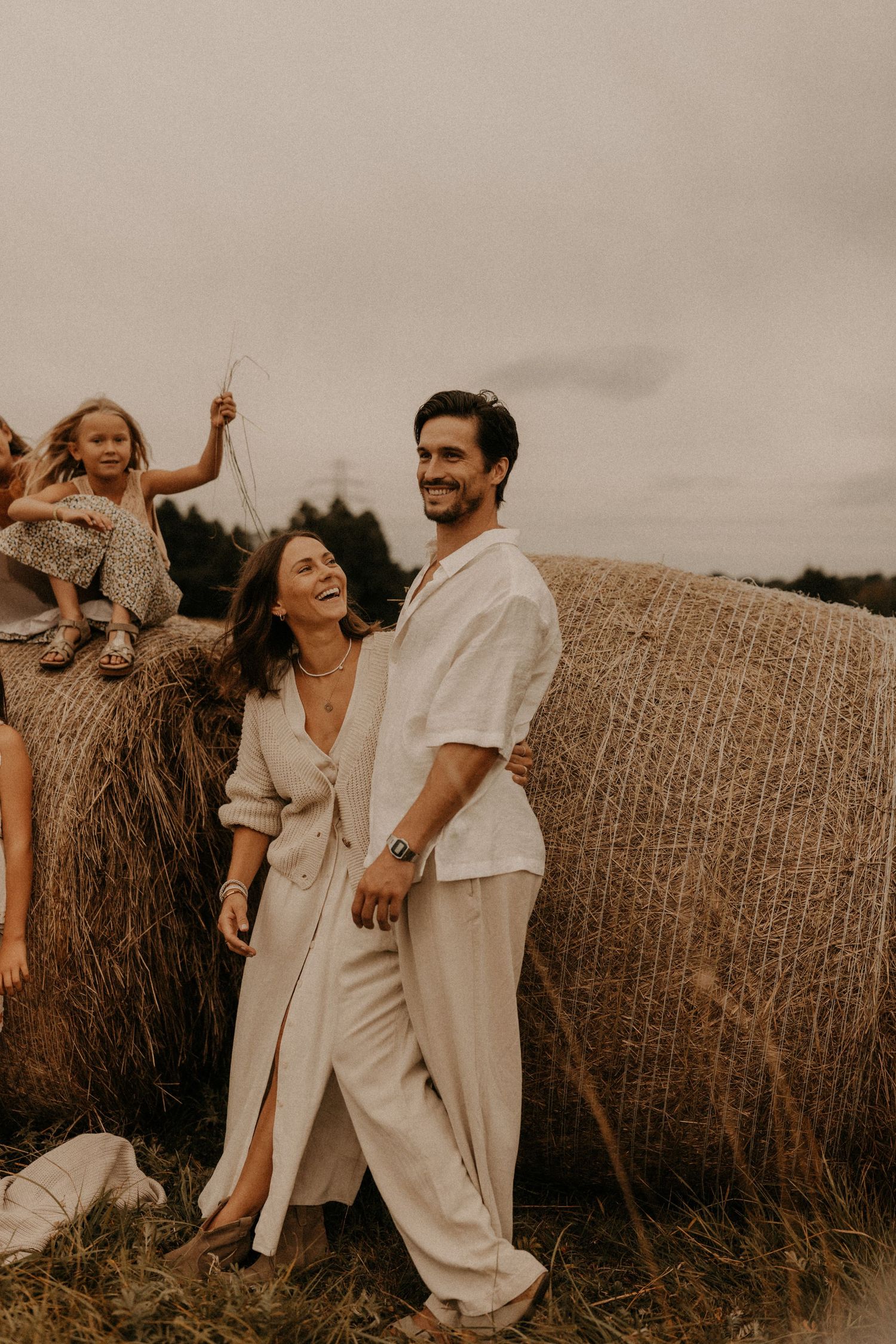 A smiling couple in neutral linen outfits poses near hay bales in a field, with a young child sitting on a bale behind them.