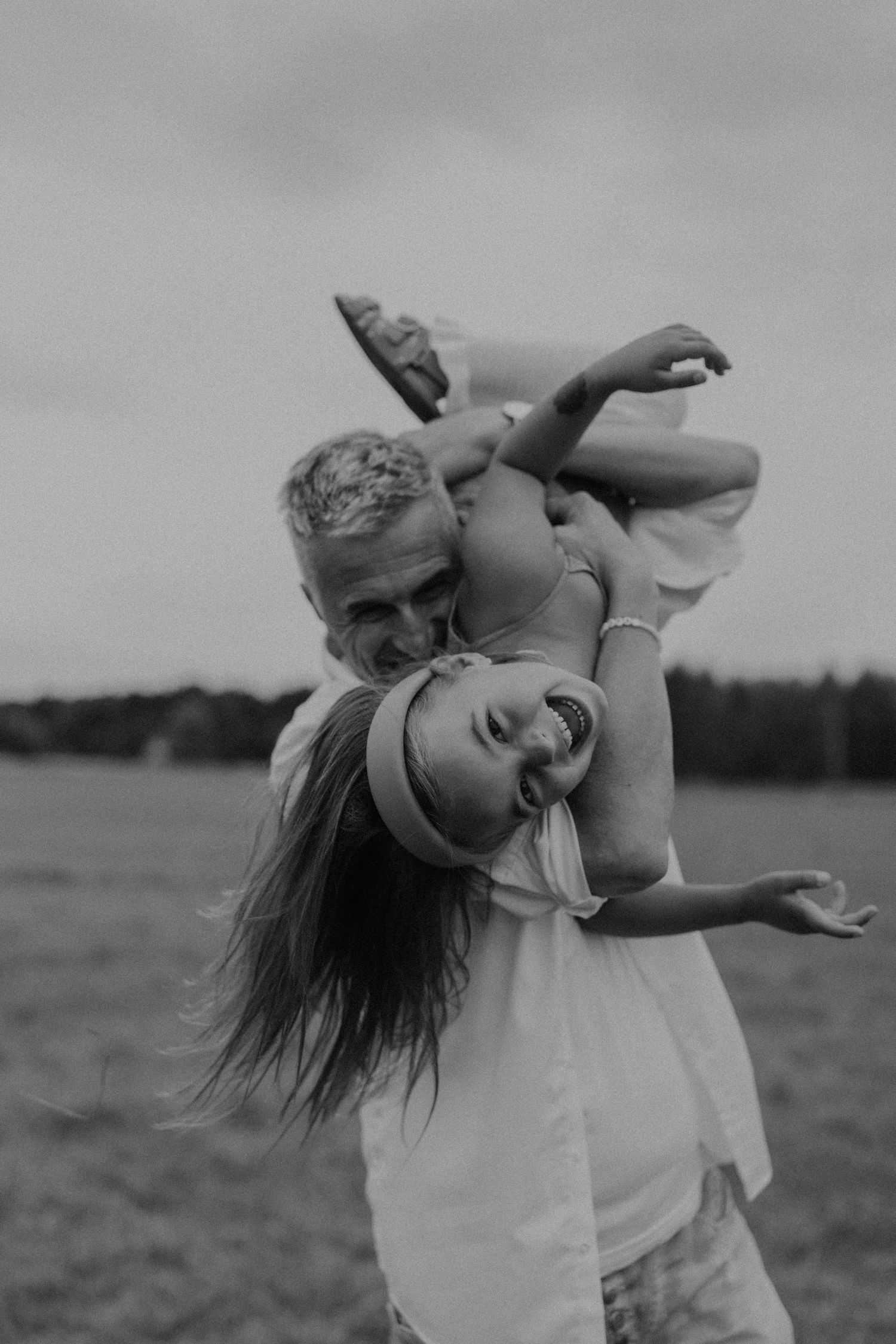 Black and white photo of a man joyfully lifting a laughing girl upside down in an open field.
