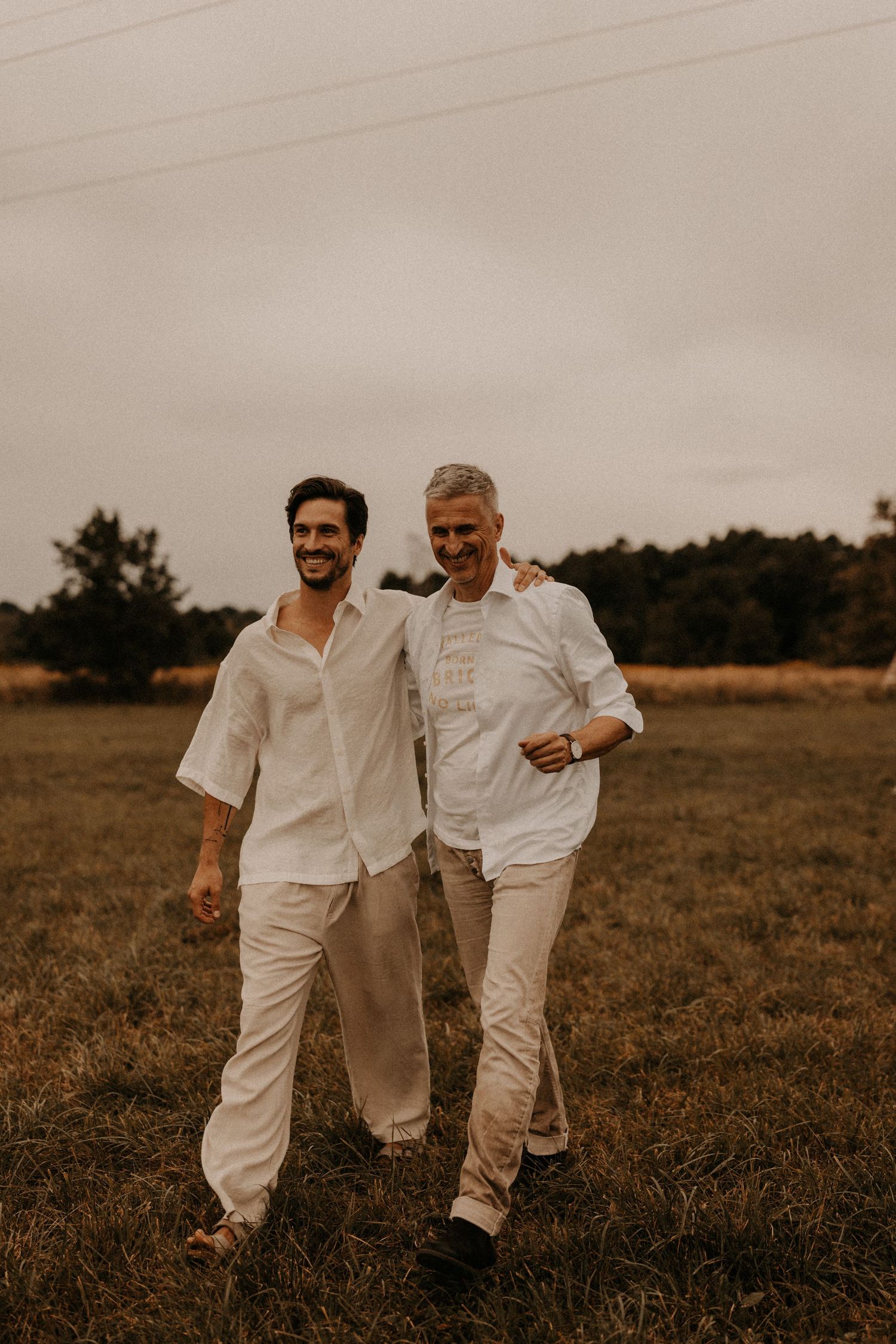 Two men in white linen outfits walking arm in arm through a green field on an overcast day.
