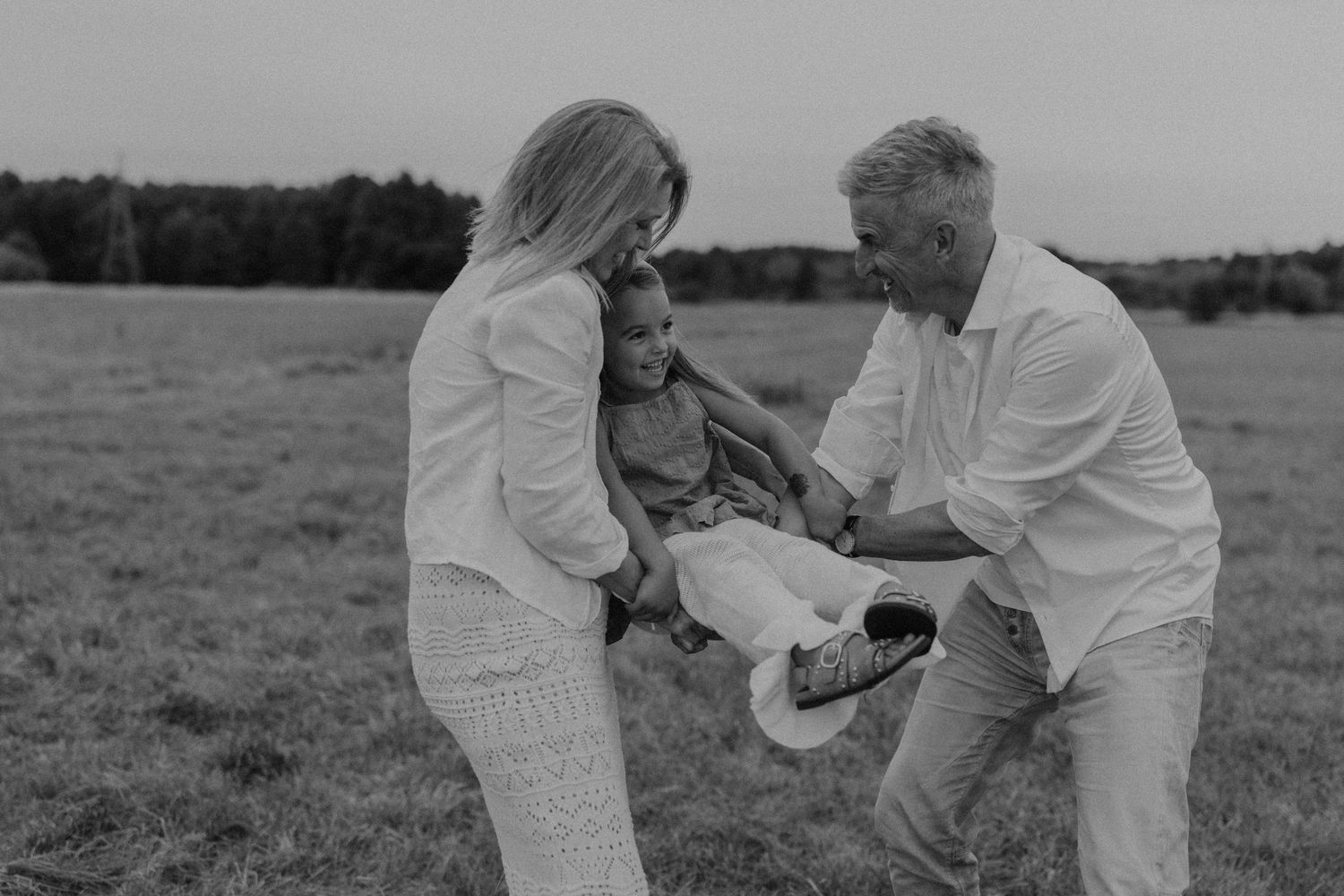 Black and white photo of grandparents playfully swinging grandchild in a field.