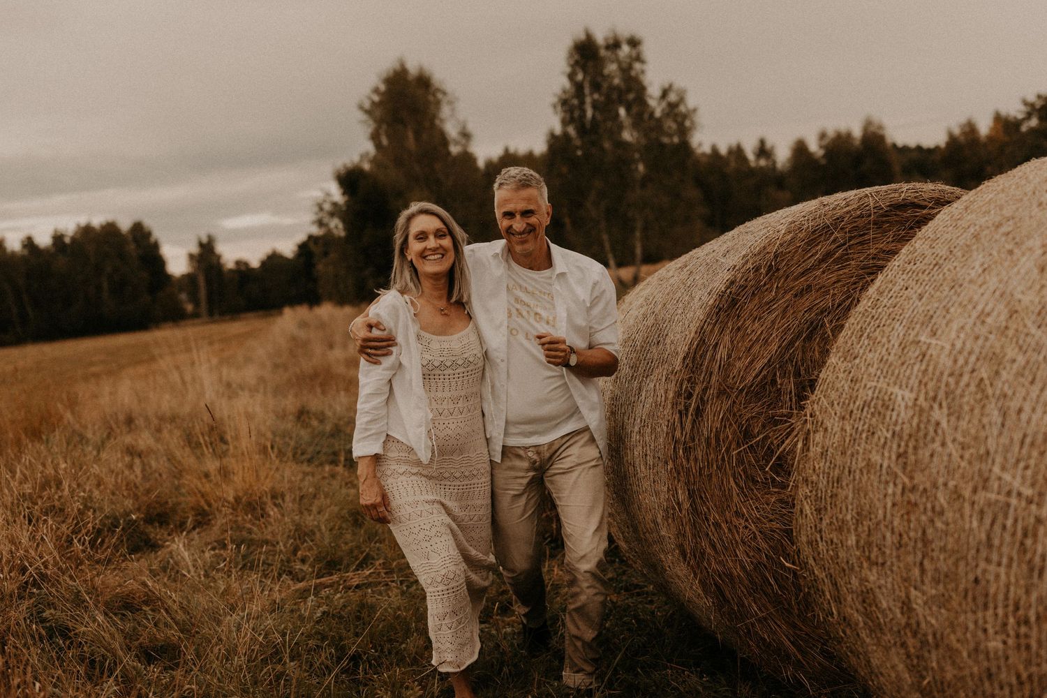 A smiling couple in white outfits walks beside a large hay bale in a golden field with forest backdrop.