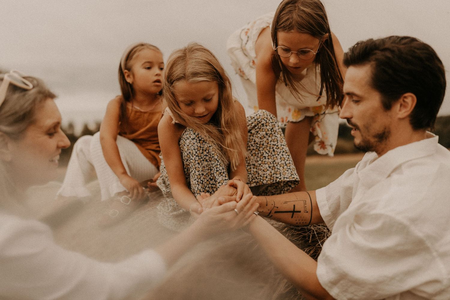 Family with three young girls gathered outdoors in soft light, holding hands in a tender, intimate moment together.