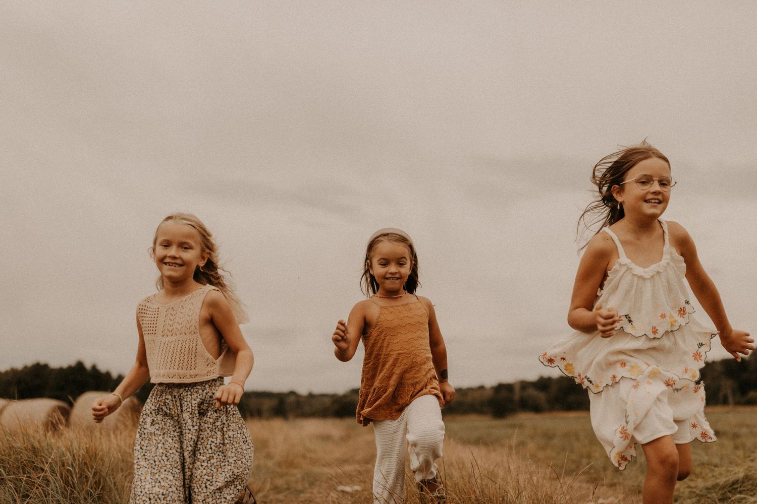 Three happy girls running through a golden field on a cloudy day, wearing boho dresses in warm tones.