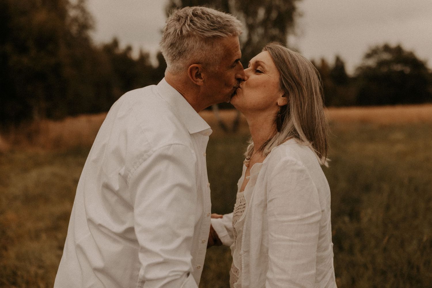 Romantic middle-aged couple sharing a tender kiss in a lush green field, both wearing matching white linen outfits.