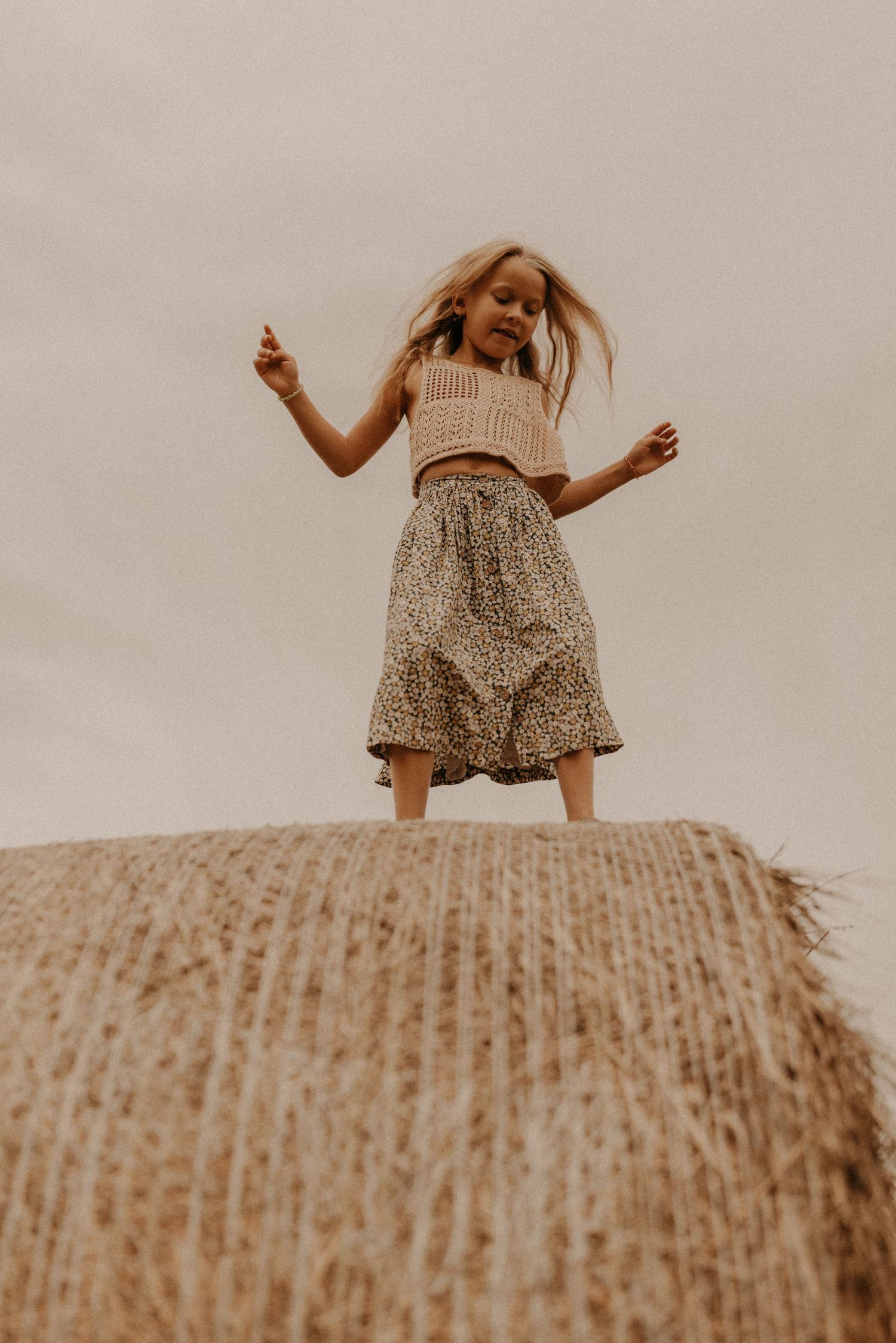 A young girl stands joyfully atop a hay bale, wearing a crop top and floral skirt, with her hair blowing in the wind.