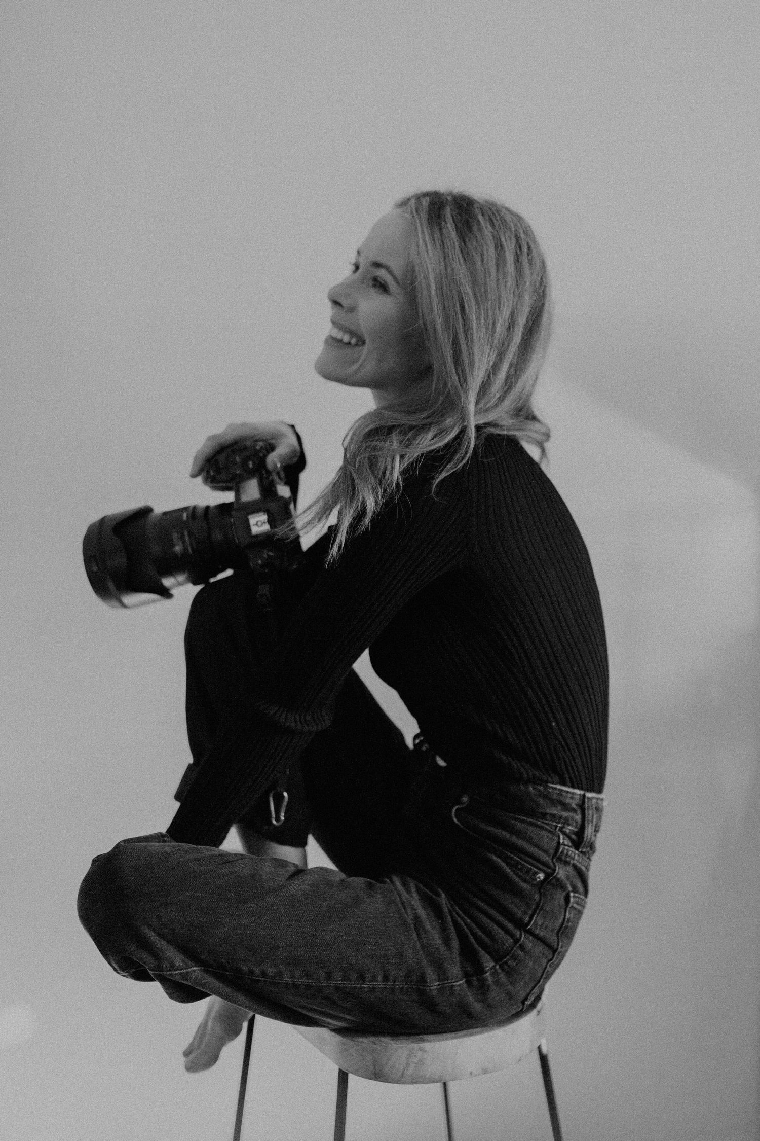 Black and white portrait of a blonde woman sitting on a stool holding a professional camera, laughing.