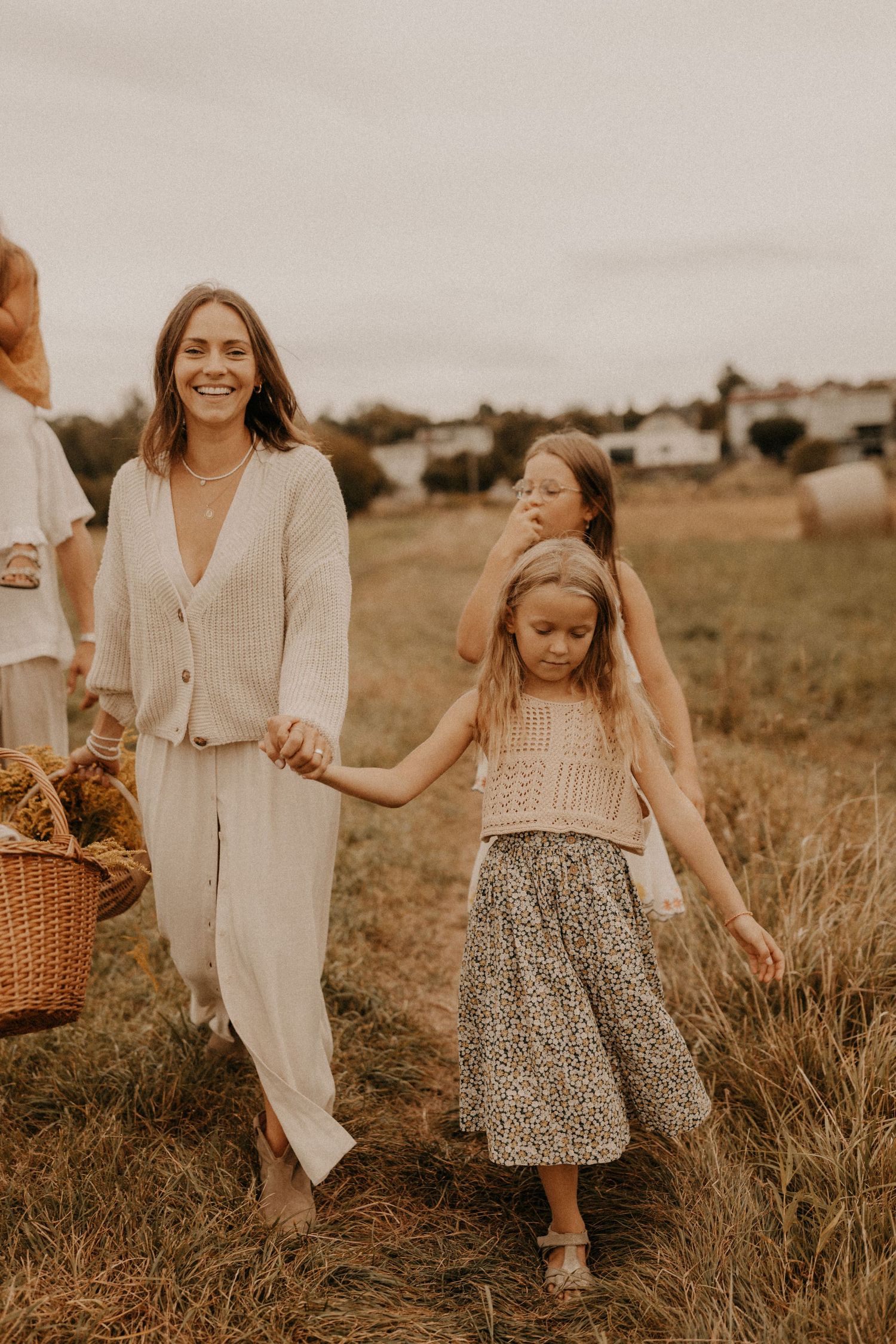A smiling woman in white linen walks through a field holding hands with two young girls carrying a wicker basket.