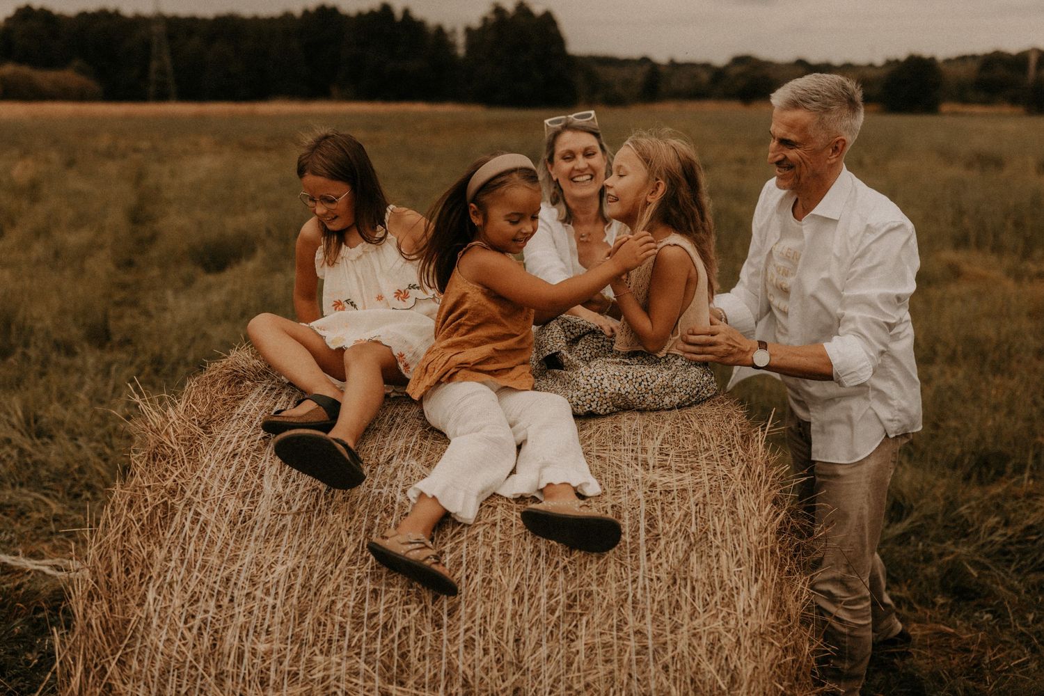 Family of five laughing together on a hay bale in a golden field during a warm outdoor photo session.