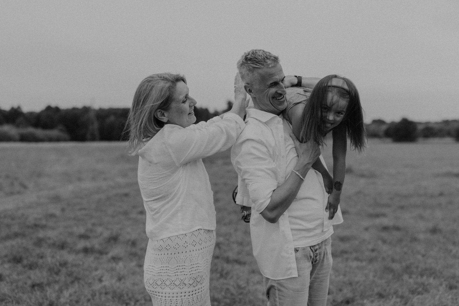 Black and white family photo of parents and daughter laughing together in an open field at dusk.