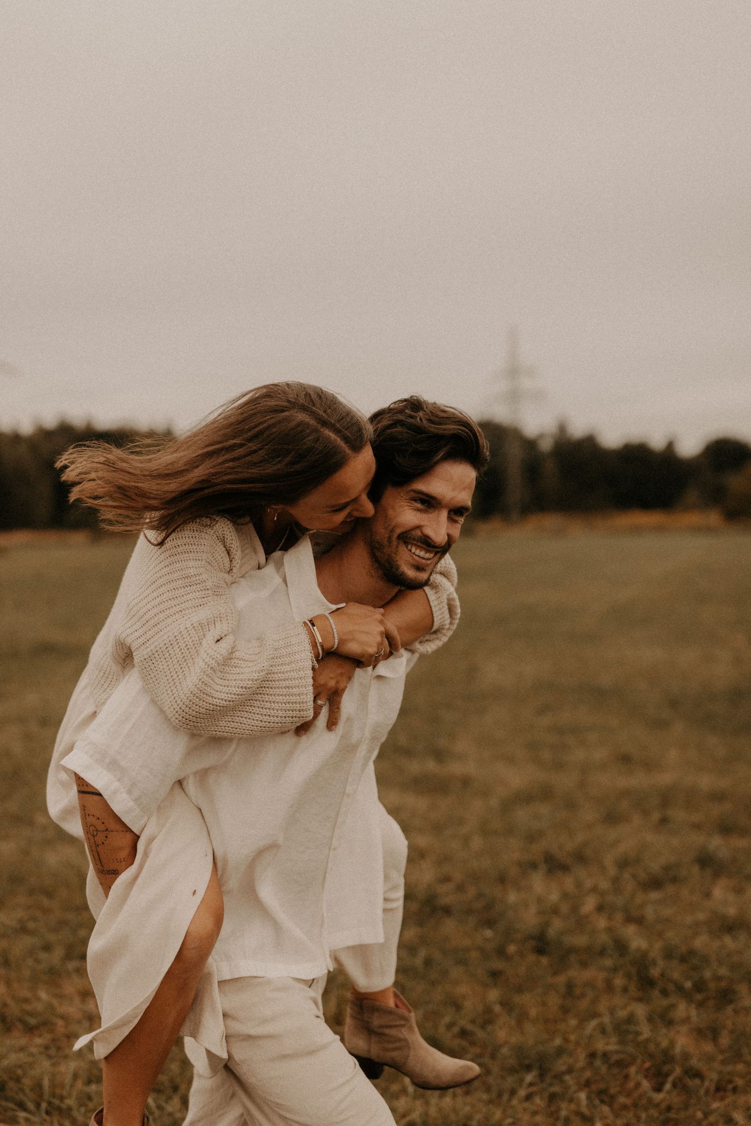A joyful couple in white outfits shares a piggyback ride in an open field, laughing together on a cloudy day.