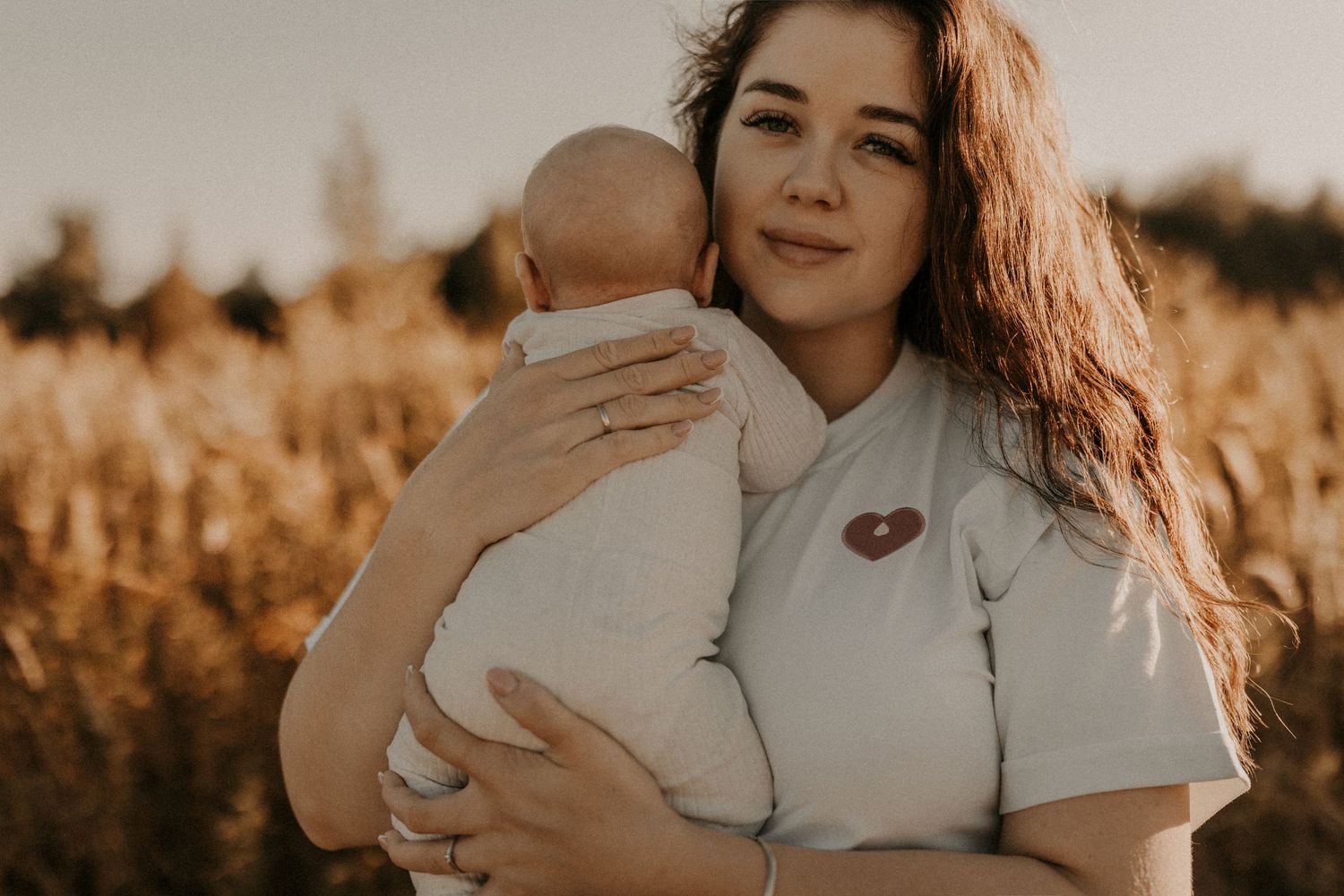 A young mother with long auburn hair holds her newborn baby in a golden wheat field at sunset.sesje zdjęciowe Rybnik, Wodzisław Śląski