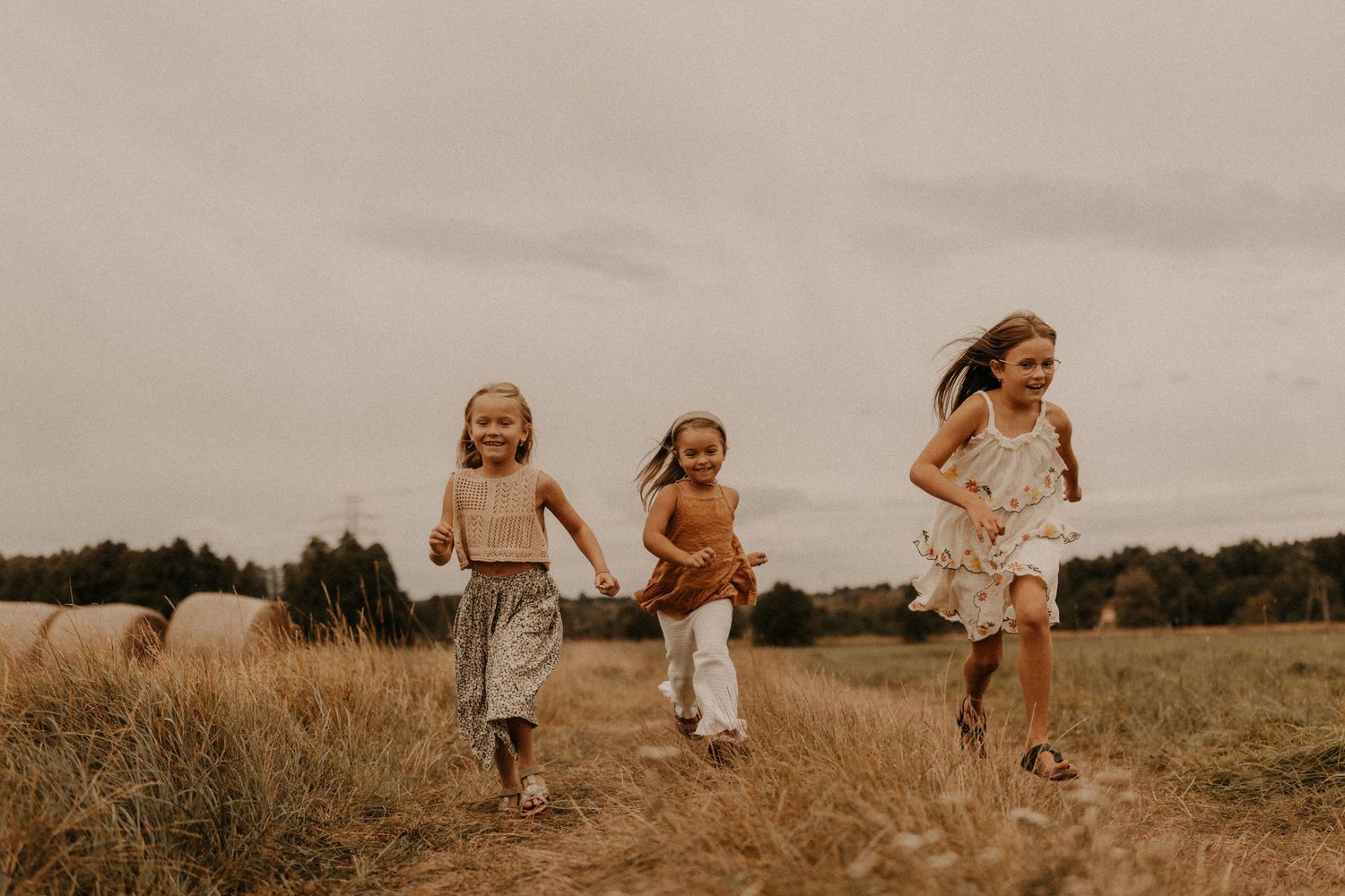 Three young girls run joyfully through a golden field on a cloudy day, wearing summer dresses in a rural outdoor setting.