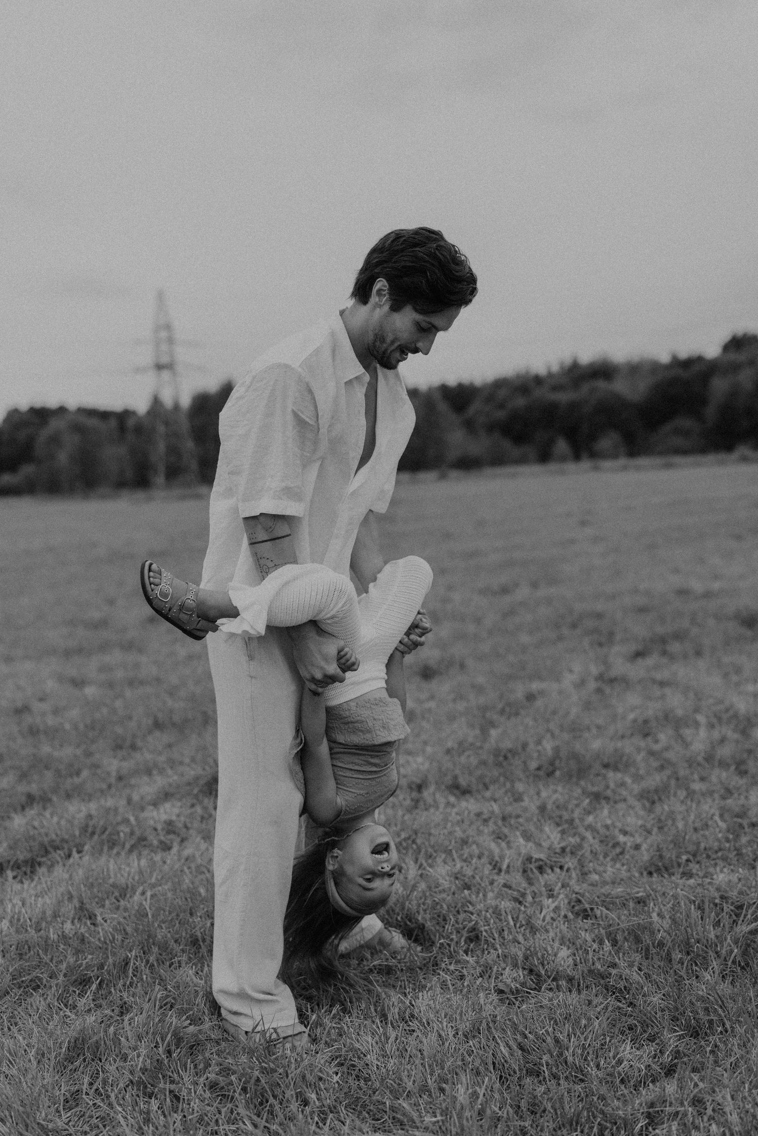 Black and white photo of a father playfully holding his child upside down in a grassy field.