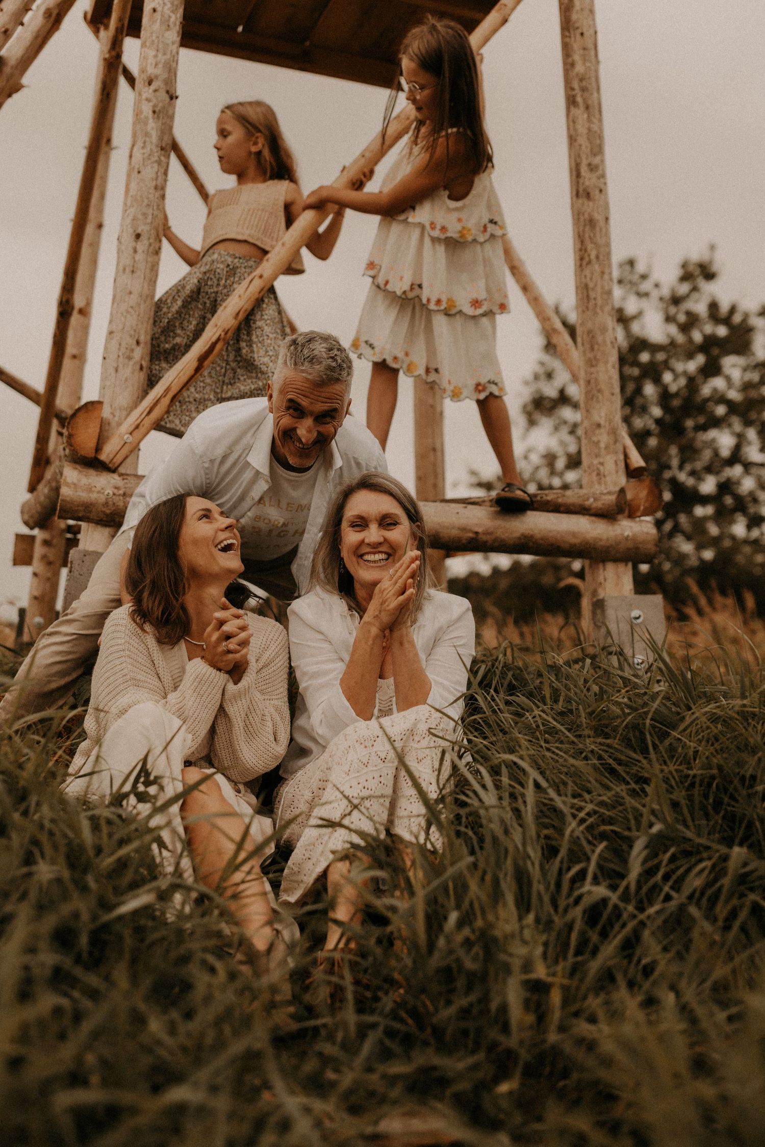A joyful multigenerational family laughing outdoors near a rustic wooden structure, with two girls playing on a ladder behind them.