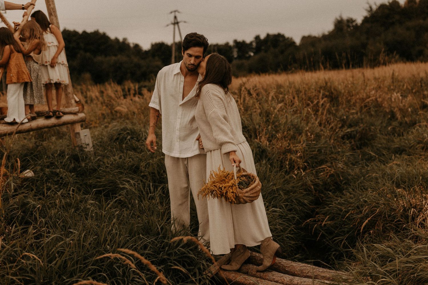 Couple in white linen outfits standing in a meadow, woman holding a wicker basket with wildflowers at dusk.
