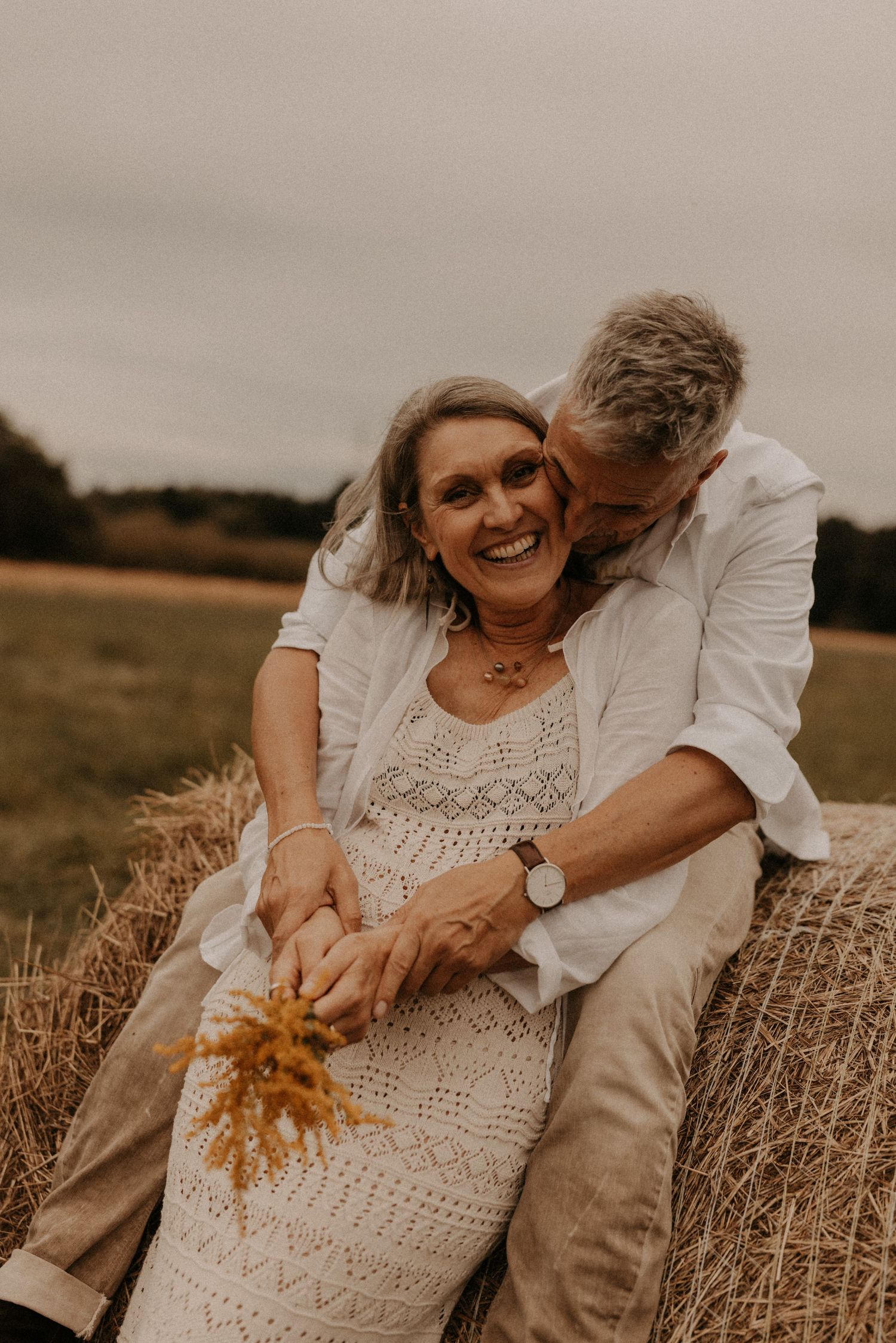 Older couple sitting on hay bale in field, woman laughing as man kisses her cheek, holding wildflowers.