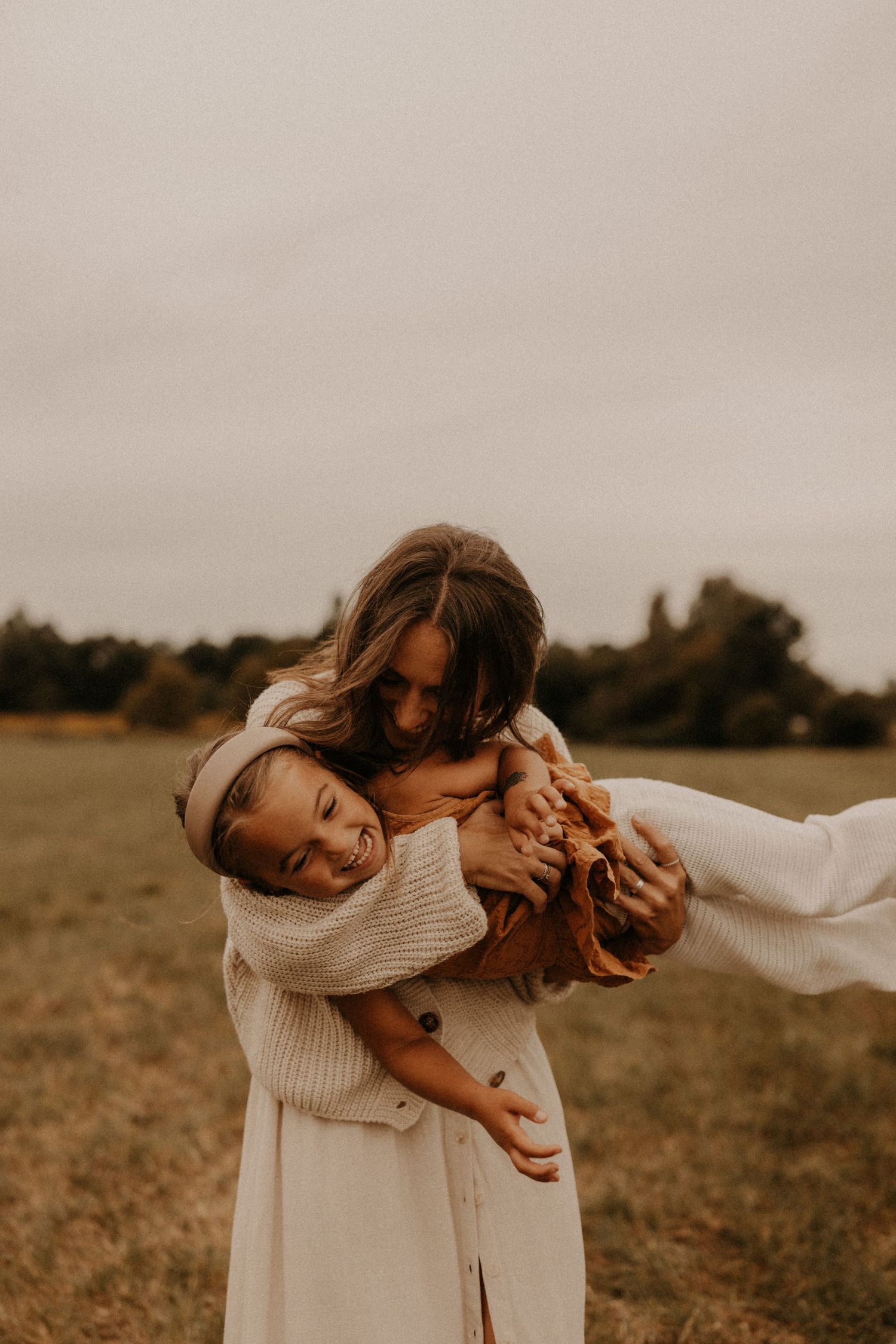 A mother joyfully embraces her laughing young daughter in an open field, both wearing cozy neutral-toned outfits.