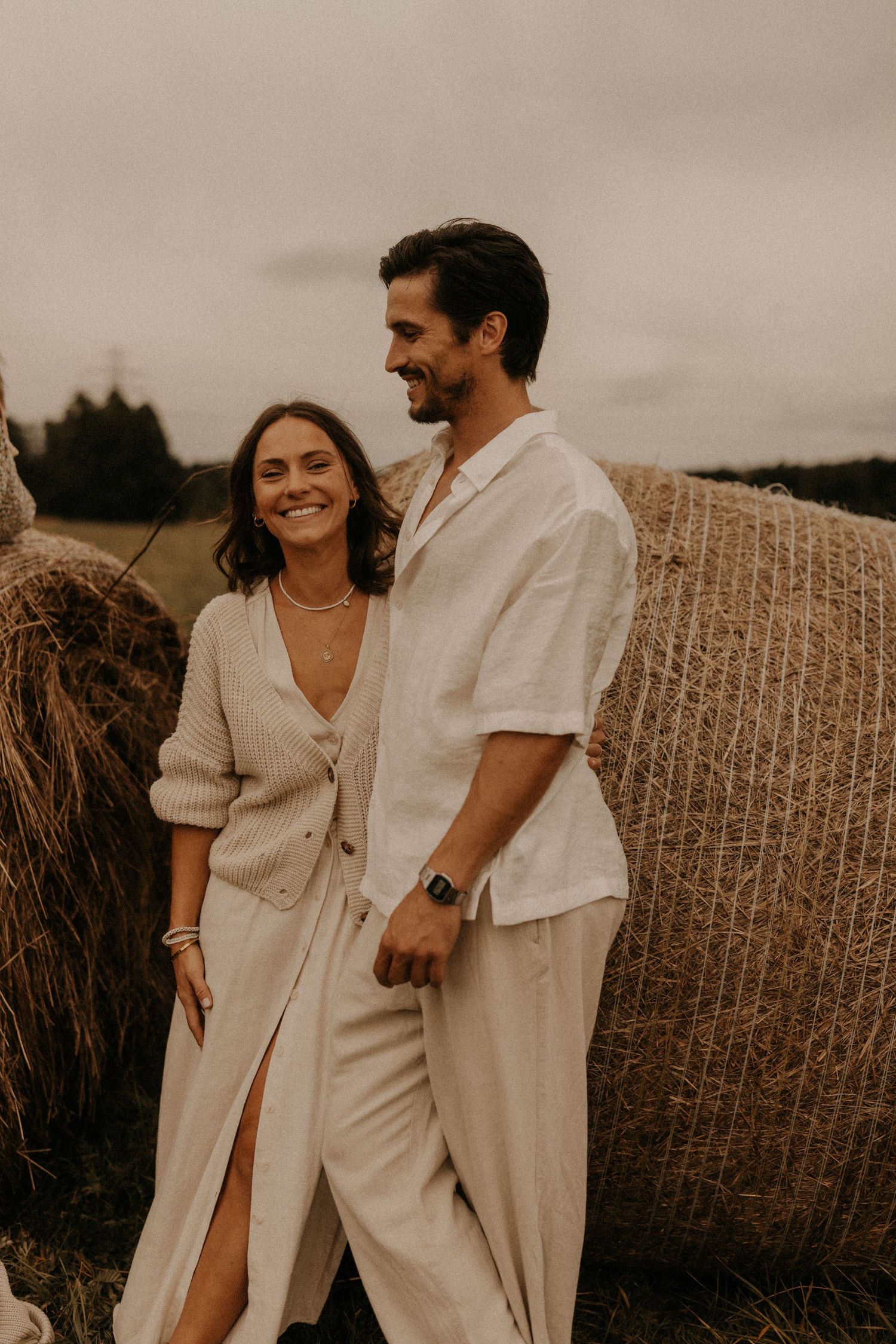 A smiling couple in neutral linen outfits poses beside hay bales in a rustic field setting.
