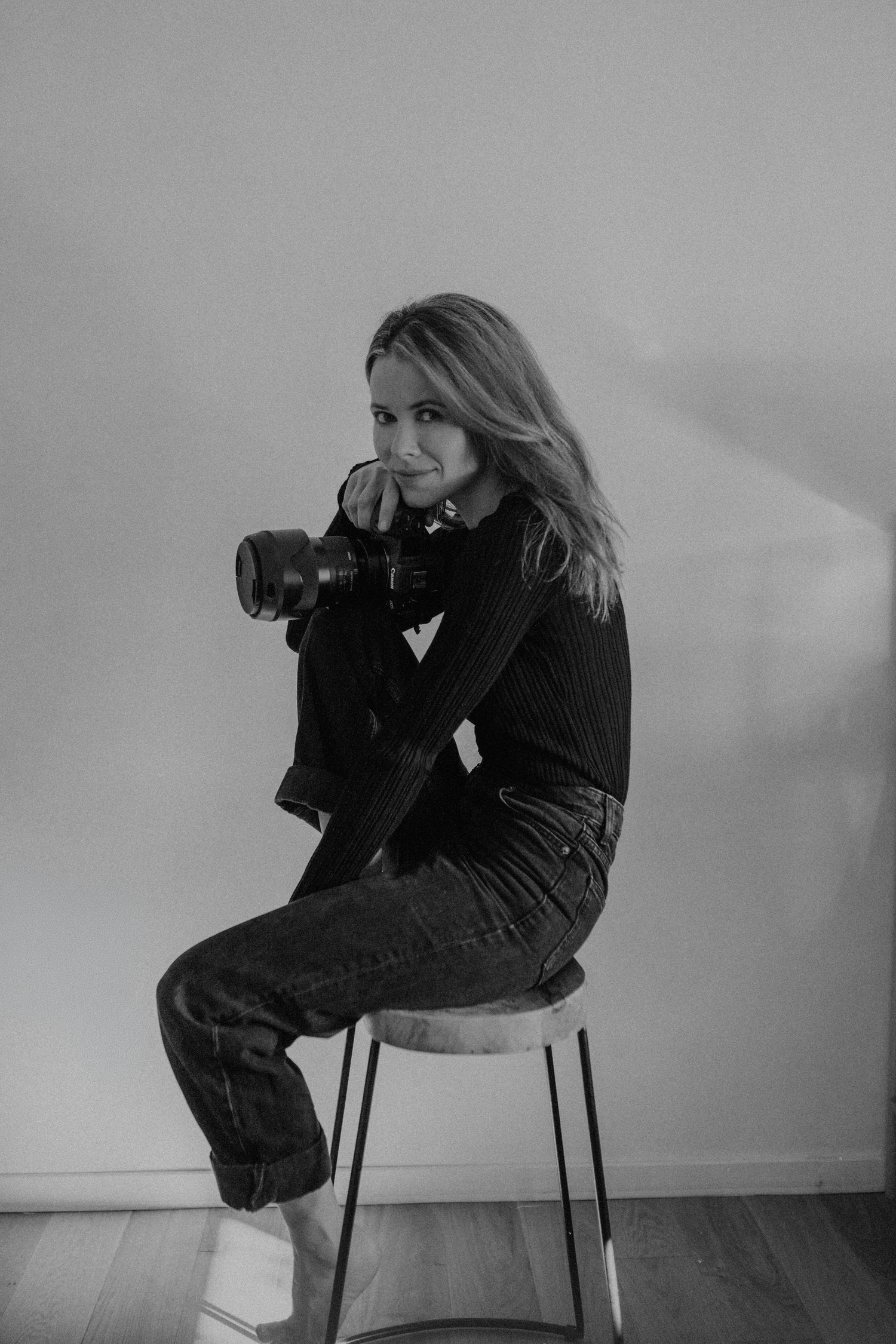 Black and white photo of a woman sitting on a stool holding a camera, wearing jeans and a dark top.esje zdjęciowe Rybnik, Wodzisław Śląski