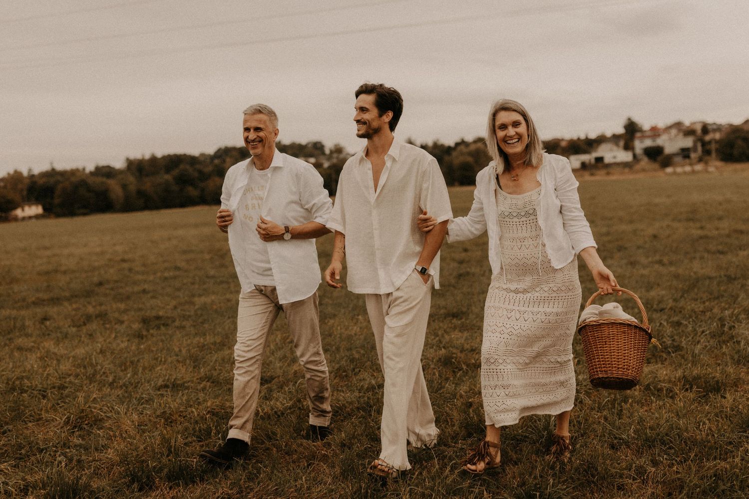 Three people in white outfits walk through a green field, one carrying a wicker basket, smiling outdoors.