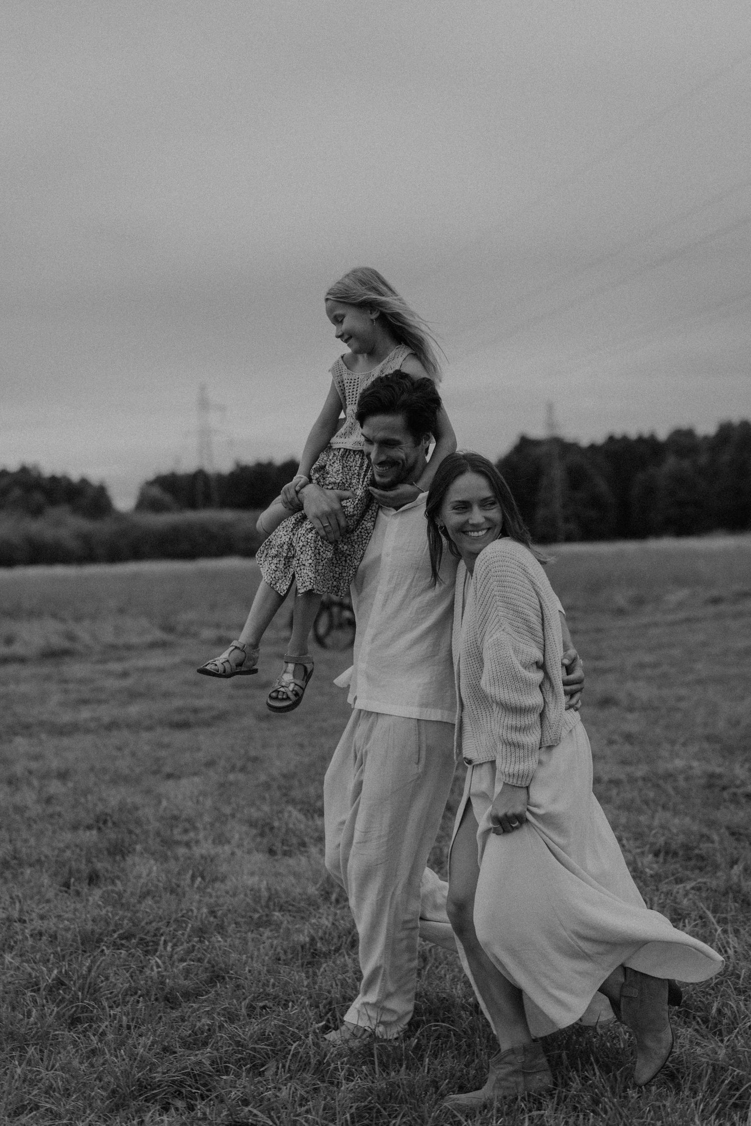 Black and white family photo of parents and young daughter on shoulders walking in a grassy field.