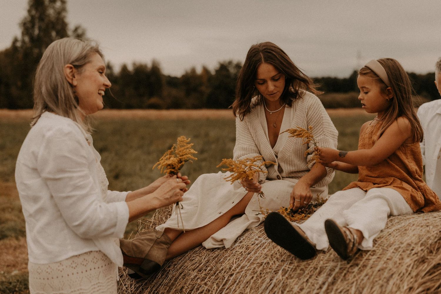Two women and a young girl sit on a hay bale in a field, sharing dried wheat stems on a warm autumn day.