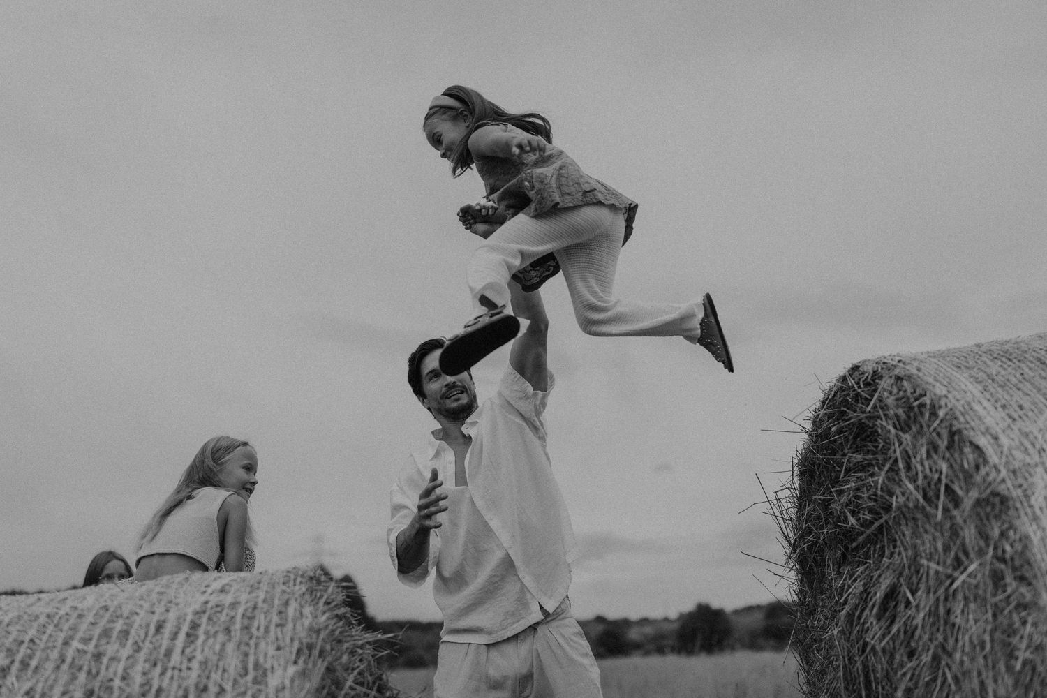 Black and white photo of a father tossing his young daughter in the air near hay bales as another child watches.