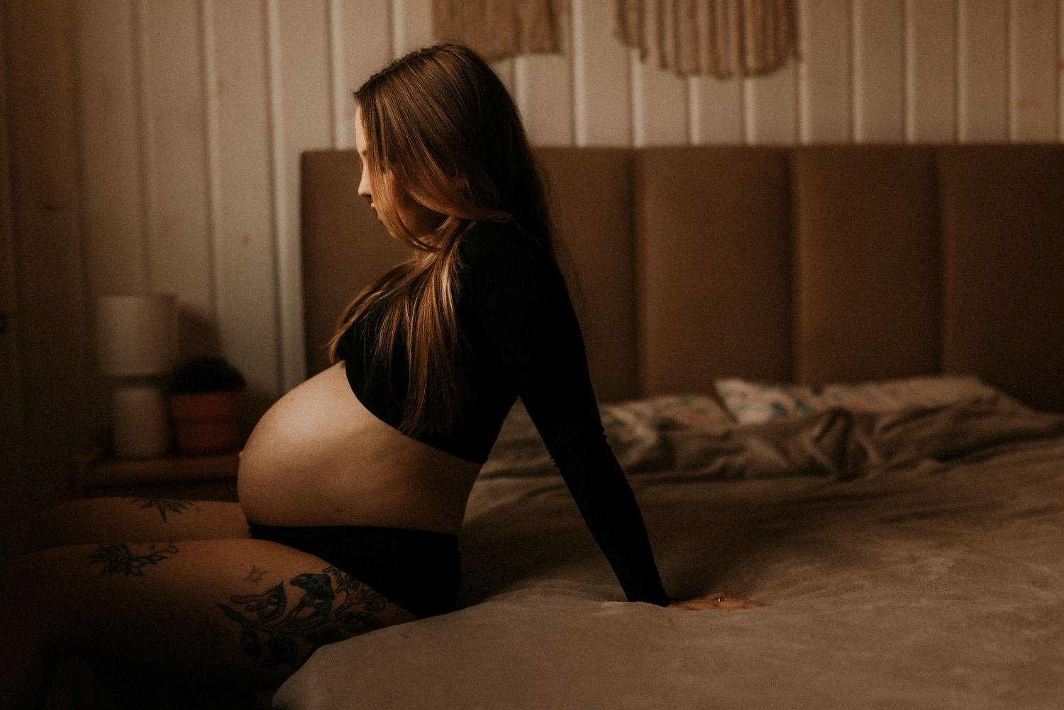 Pregnant woman in black outfit sitting on bed, showcasing baby bump in moody bedroom maternity photo. fotograf Rybnik, Wodzisław Śląski