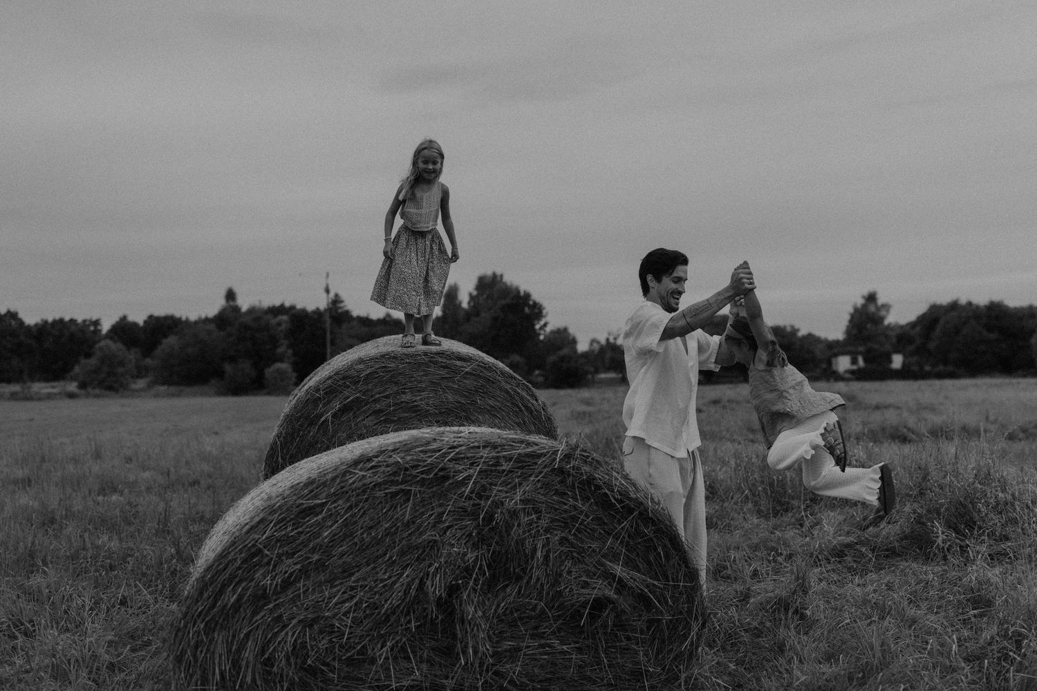 Black and white photo of children playing on hay bales in a field under a cloudy sky.