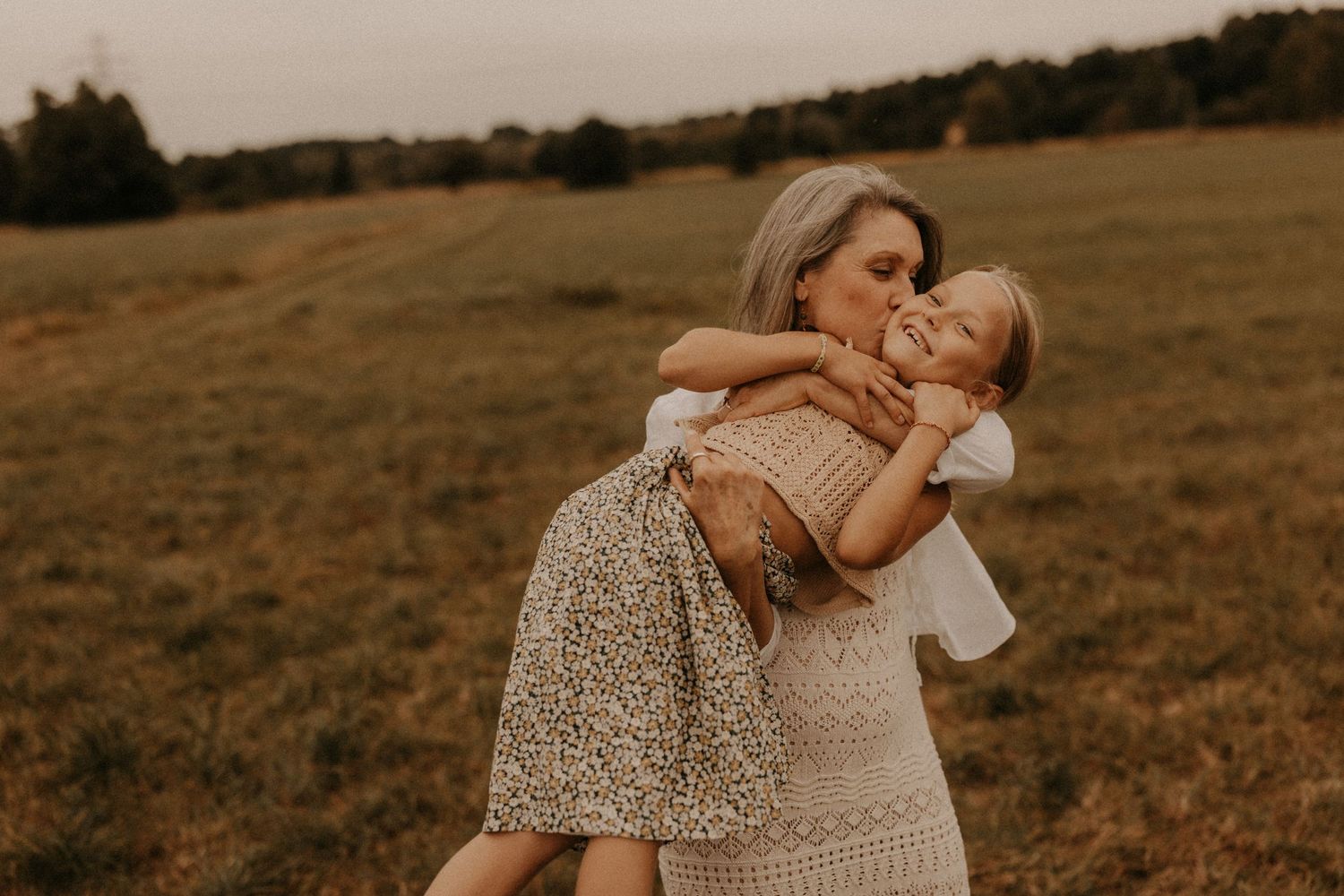 A mother joyfully embraces her young child in a sunlit green meadow, both wearing light floral outfits.