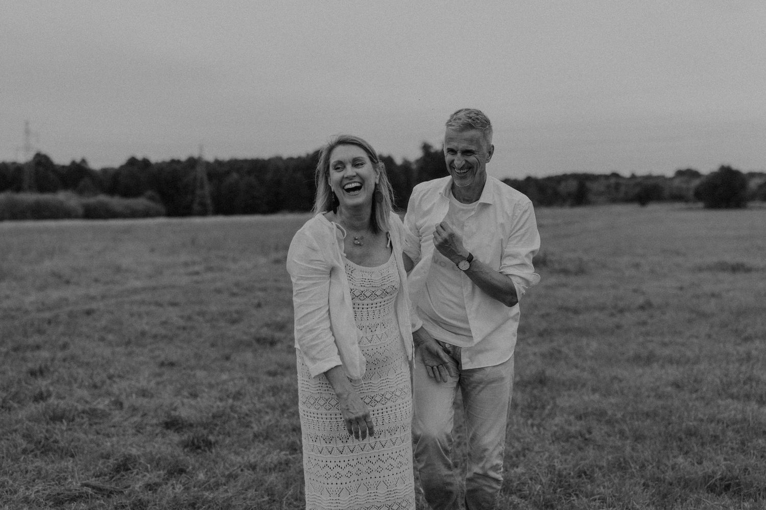 A laughing couple walks together in a field, the woman in a white lace dress and man in casual white attire.