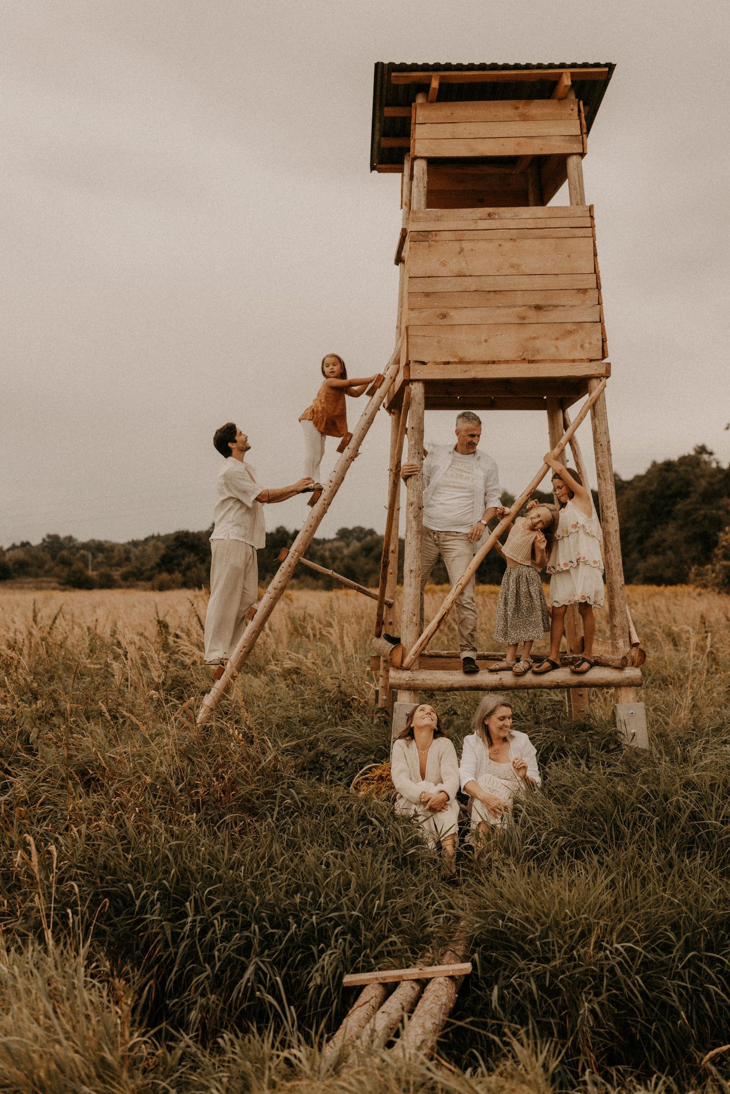 Family gathering around a rustic wooden watchtower in a golden field, dressed in white and earthy tones.