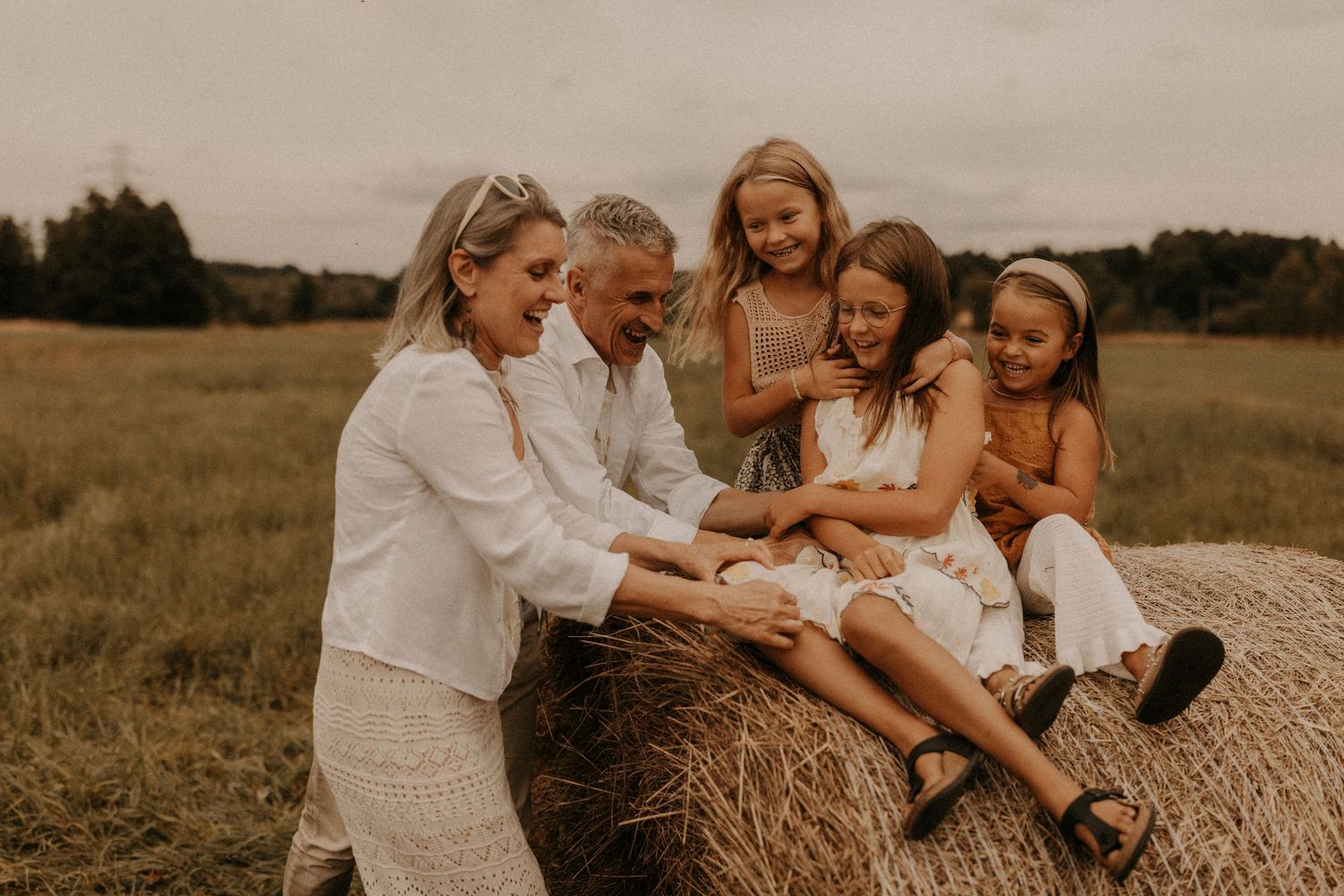 Family laughing together on a hay bale in a field, with two adults and three girls enjoying a sunny outdoor moment.