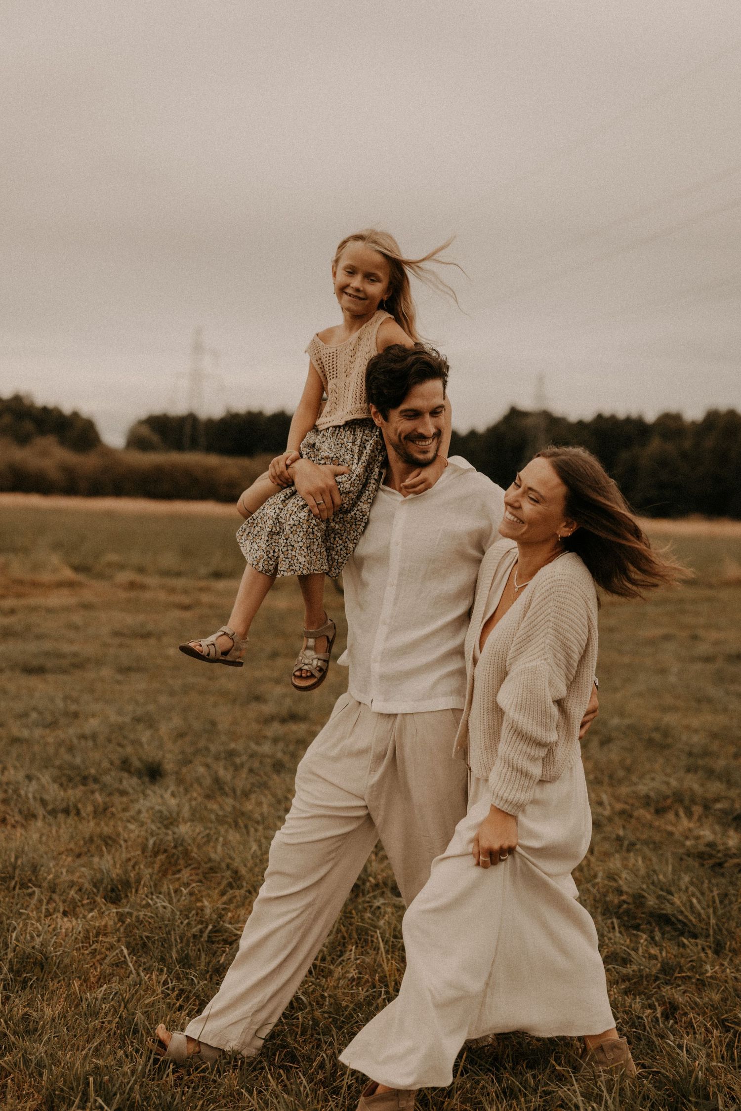 A joyful family of three walks through a field, with a man carrying a laughing girl on his shoulders beside a smiling woman.