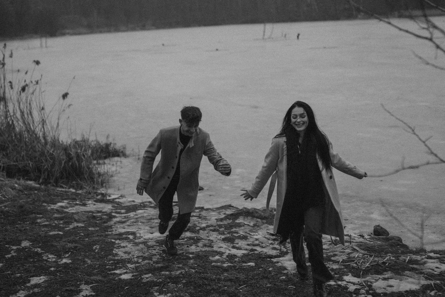 Black and white photo of a couple running and laughing together near a frozen lake outdoors.sesje zdjęciowe Rybnik, Wodzisław Śląski