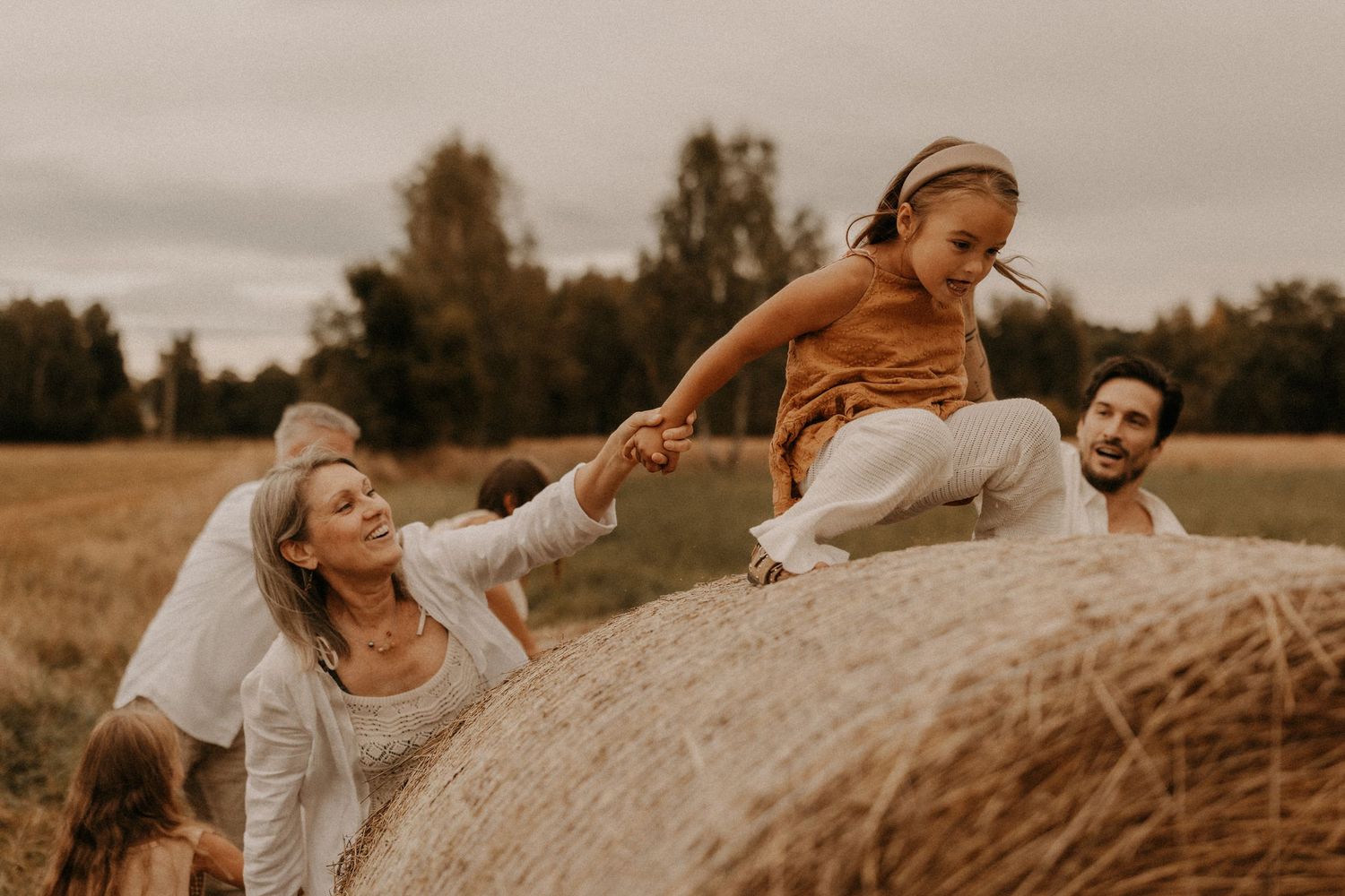 Family playing on a hay bale in a field, with a young girl climbing up while parents laugh outdoors.