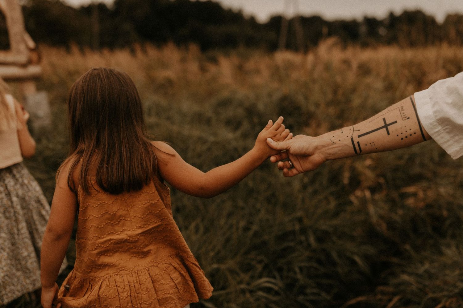 A young girl in an orange dress holds hands with a tattooed adult in a golden field at dusk.