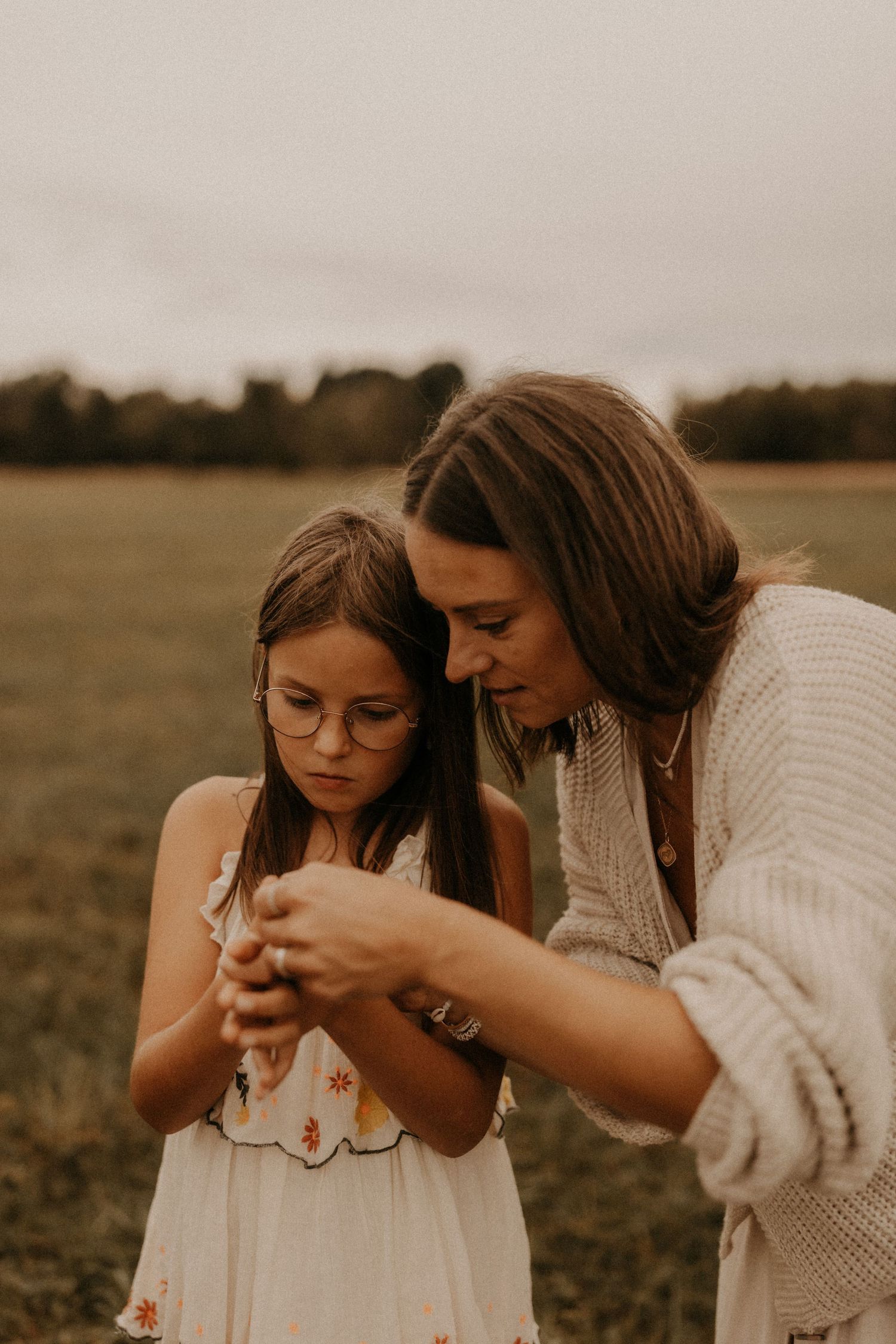 A woman and young girl with glasses look down at something in their hands in a green field.