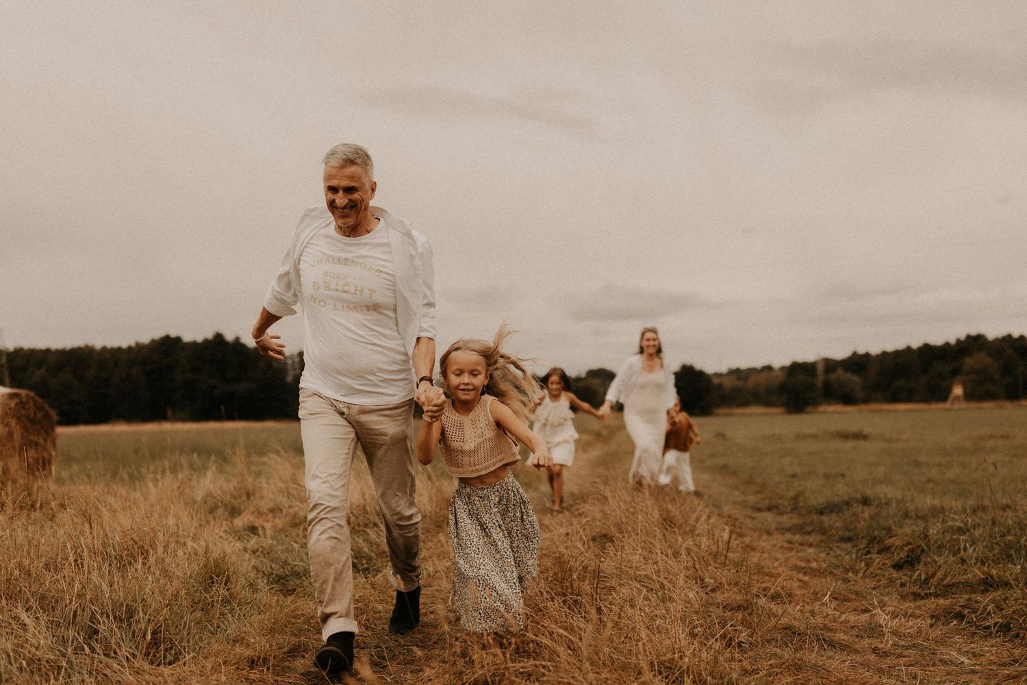 Family running joyfully through a golden field, grandparents and children enjoying outdoor time together on a cloudy day.