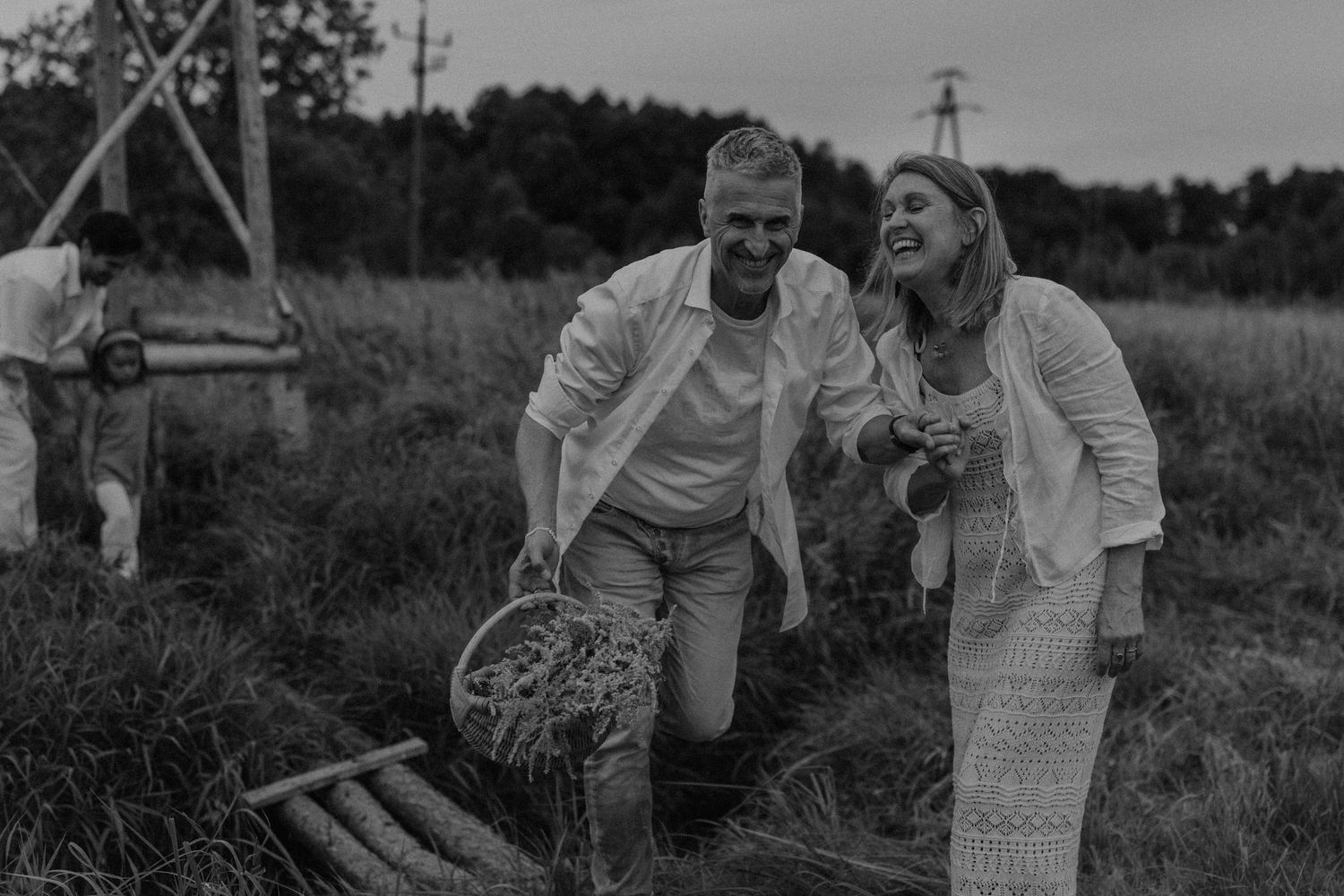 Black and white photo of a smiling couple walking through a field, man carrying a basket, with a person working in the background.