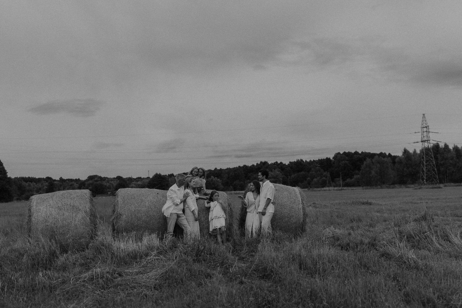 Black and white photo of a family gathering around hay bales in a rural field with trees and a power line in the background.