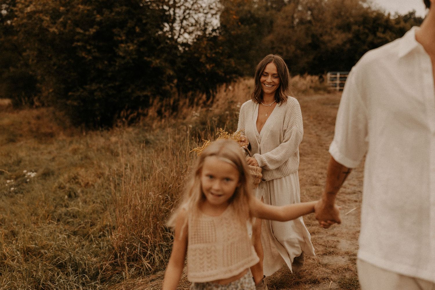 A smiling woman in a white dress walks with a young blonde girl holding wildflowers in a sunlit field.