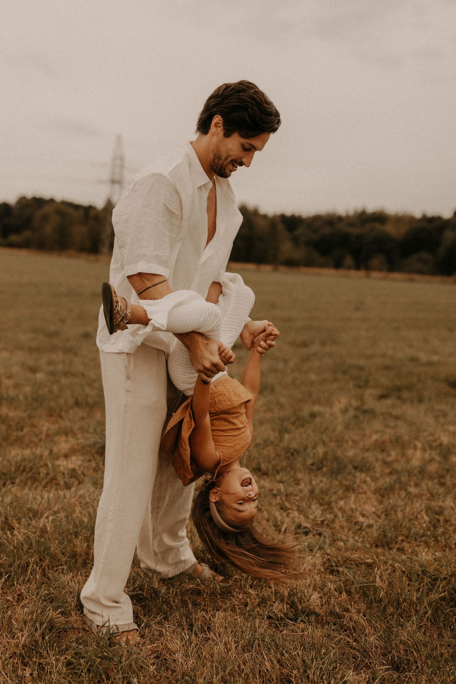 A smiling man in white linen playfully holds a laughing child upside down in a green field.