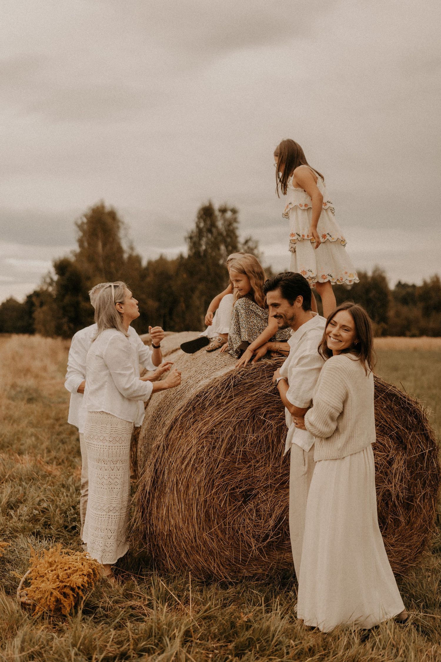 Family in white outfits posing around a large hay bale in an open field during an autumn photo session.