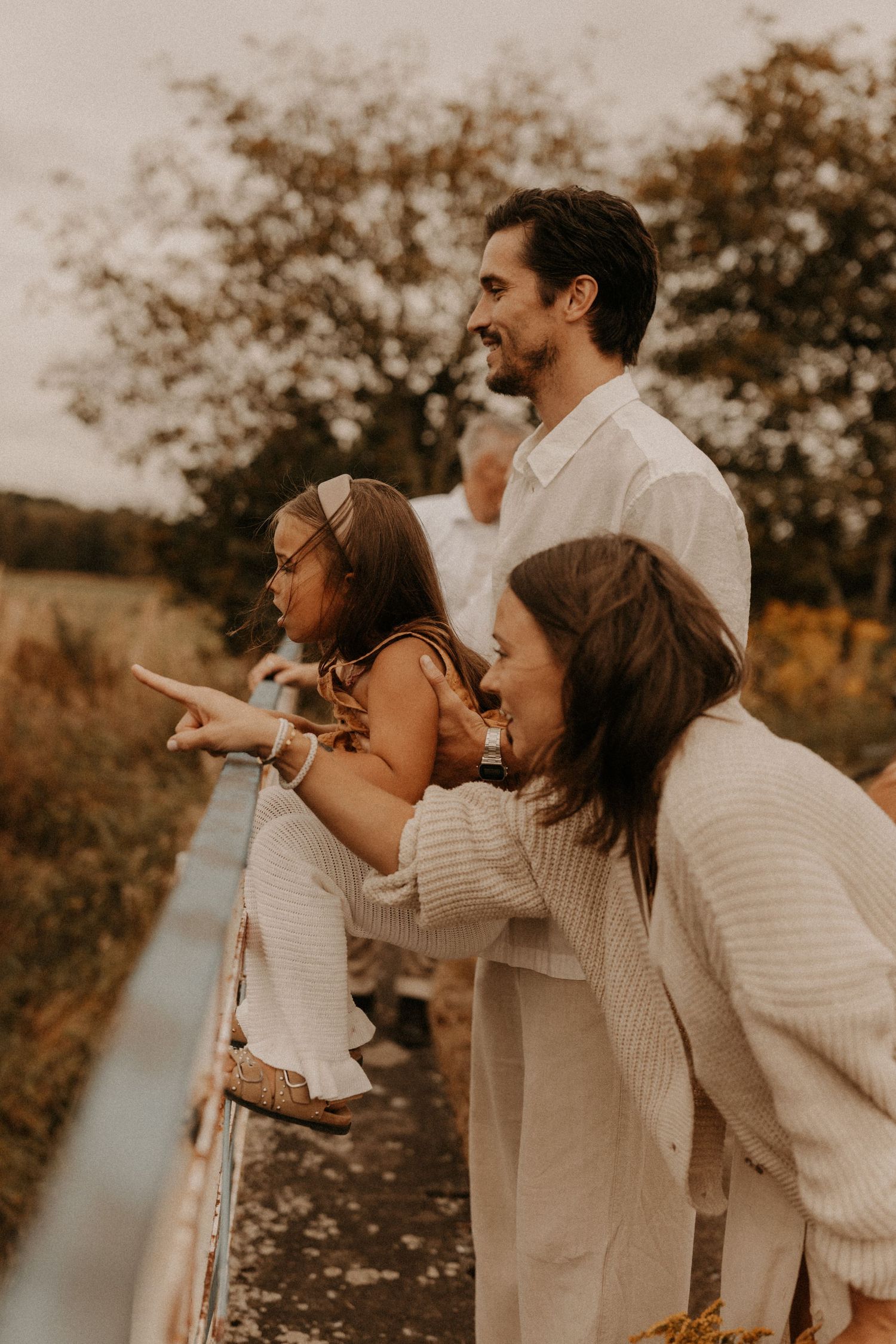 Family in cream outfits standing on a bridge, smiling and pointing at a scenic outdoor landscape.