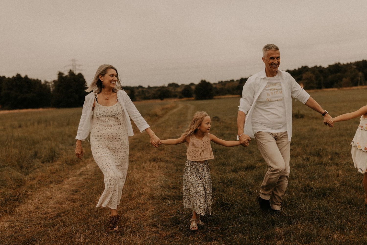 Family of three holding hands walking through a green field, parents and young daughter in neutral tones.