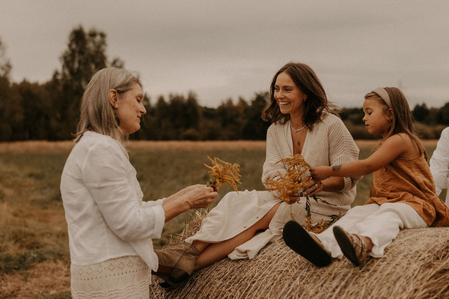 Three women sit on a hay bale in a field, sharing wildflowers and smiling on a cloudy autumn day.