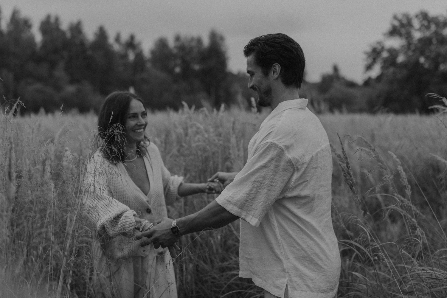 Black and white photo of a couple holding hands and smiling in a tall grass field with trees in background.