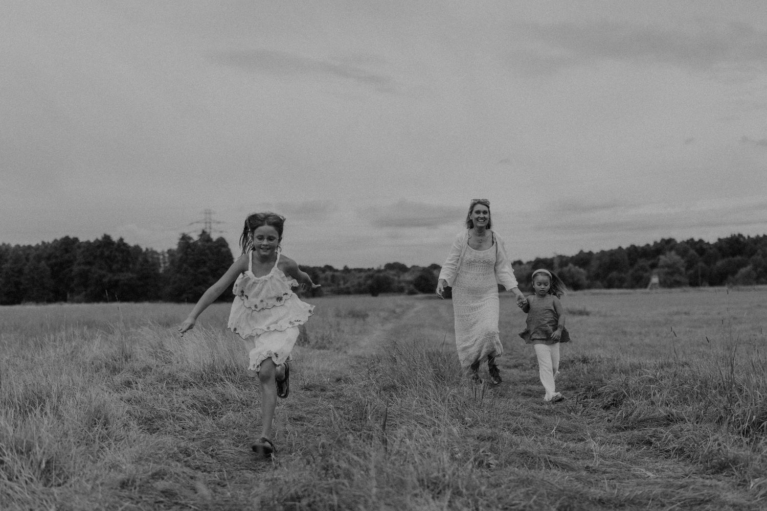 Black and white photo of a woman and two children running joyfully through a grassy field with trees in the background.
